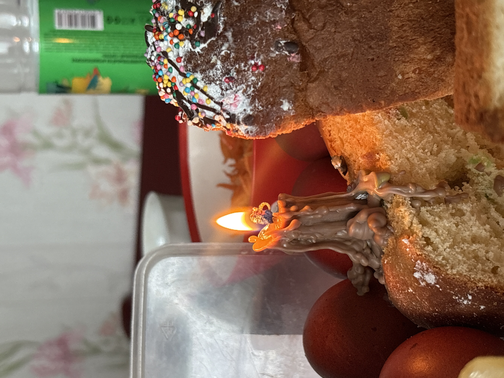 A close-up, high-angle shot depicts a section of Easter bread, likely a Kulich, adorned with colorful sprinkles on top. The bread is partially cut, revealing its interior. A lit candle with elaborate, melted wax flows down the side of the bread. Several red-brown dyed Easter eggs are situated beneath the bread. The setting appears to be indoors, possibly a kitchen or dining area. A bottle, a floral-patterned surface, and a portion of green packaging are visible in the background. The specific location is Пожарское сельское поселение, Ukraine.