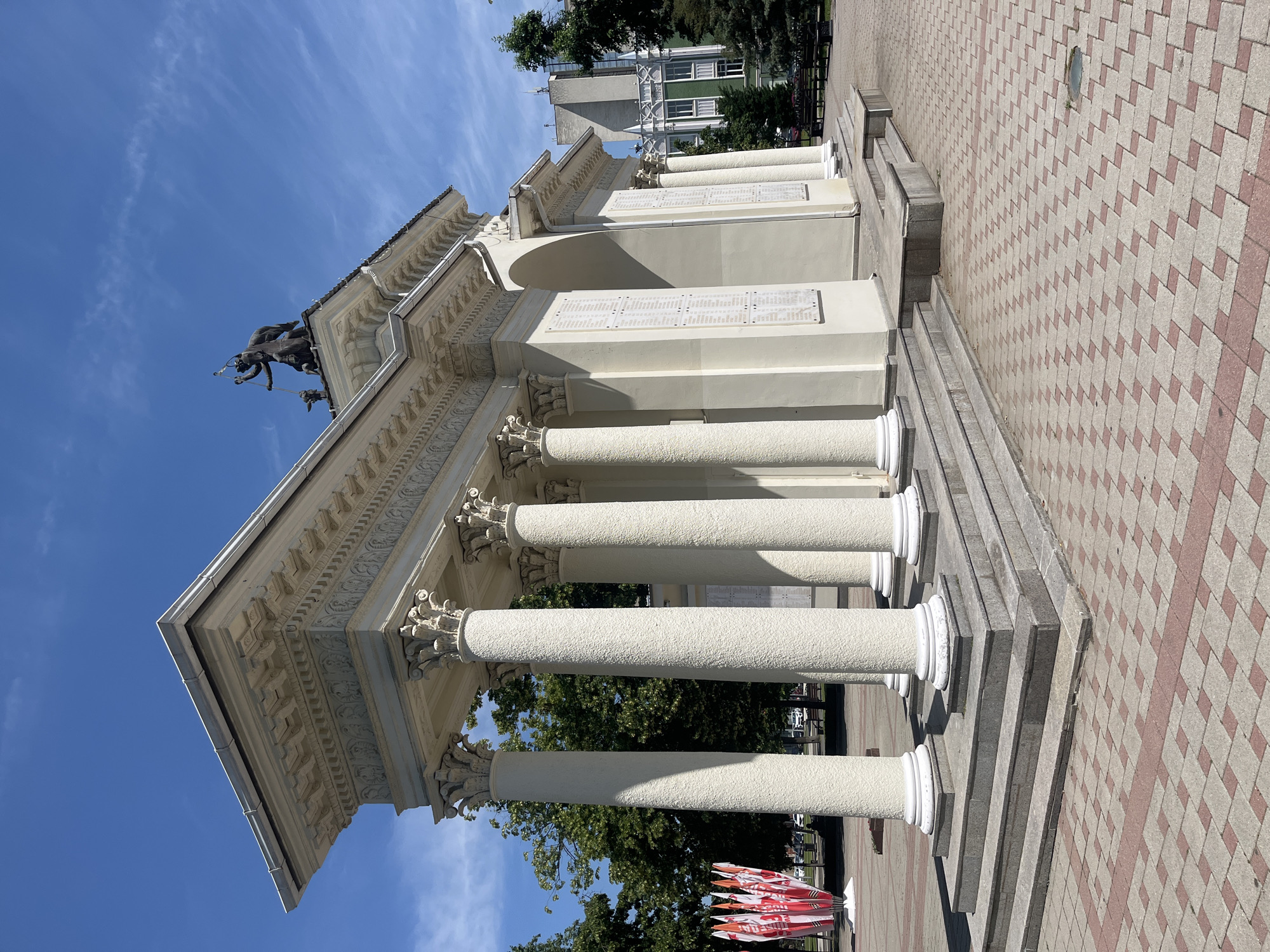 The image shows the Arch of Victory in Krasnodar, Russia. The arch is a white, neoclassical structure with a portico of Corinthian columns and an entablature with decorative carvings. Atop the arch is a sculpture of a winged goddess and a soldier. The arch is set on a paved plaza with red and gray tiles. In the background, there are trees and modern buildings, and a display of red and white flags. The sky is blue with some scattered clouds. The image is taken on a sunny day.