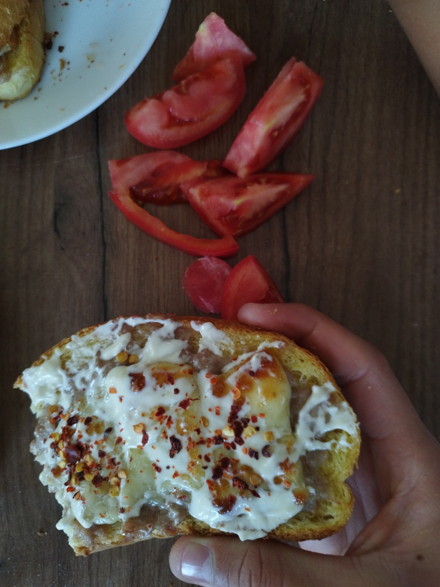 A close-up shot depicts a half-eaten slice of bread held in a human hand, topped with melted cheese, sauce, and red pepper flakes. Adjacent to the bread are several slices of fresh tomato, scattered on a wooden surface. A white plate with remnants of food is partially visible in the upper left corner of the frame. The image suggests a snack or light meal being prepared or consumed indoors. The location is unknown.