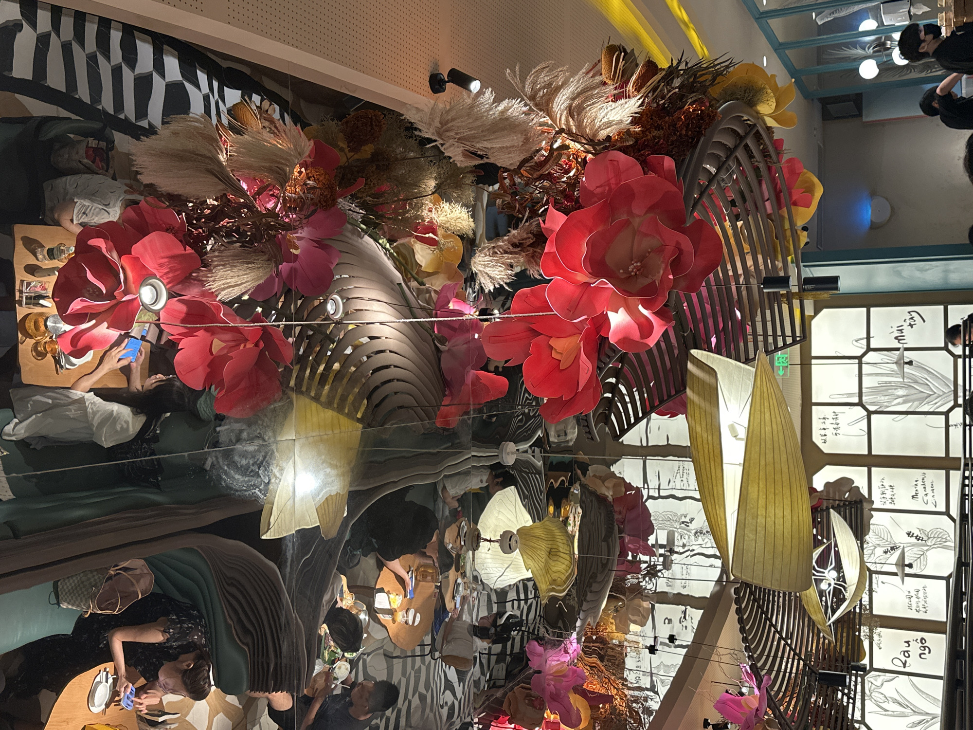 The image depicts a restaurant interior, likely in Futian District, China, characterized by its reflective ceiling and elaborate floral and decorative installations. The ceiling is highly polished, mirroring the scene below and creating an illusion of depth and an upside-down perspective. Dominating the view are oversized, vibrant red and pink paper flowers, interspersed with feathery dried grasses and other textured botanical elements, arranged in cascading floral arrangements.
Below these decorations, reflections reveal snippets of the restaurant's dining area. Several tables are visible, some occupied by people. In one reflection, a woman wearing a dark patterned dress is seated at a table, holding a blue mobile phone. Another reflection shows individuals seated around round tables, with drinks and food items present. In the foreground of the main image, and reflected in the ceiling, are unique pendant lamps that appear to be crafted from woven material, emitting a warm yellow light.
The walls visible in the reflections are decorated with large-scale botanical illustrations and text, some of which appear to be in Asian script. The overall ambiance is one of contemporary, artistic dining, with a focus on visually striking decor. The lighting is a mix of spotlights illuminating the floral arrangements and the ambient light from the lamps. The overall impression is one of a sophisticated and visually engaging dining space.