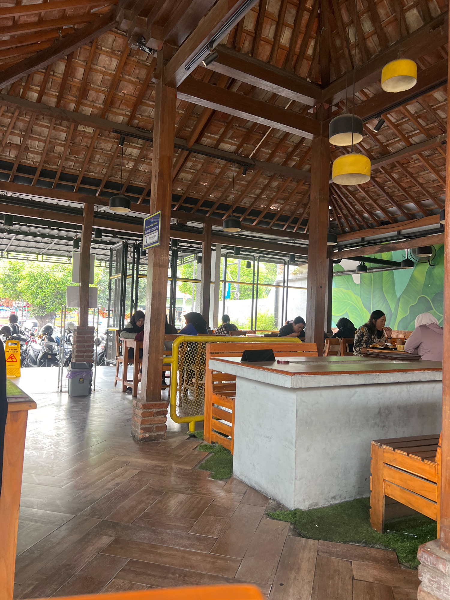 The image depicts the interior of a cafe or restaurant in Yogyakarta, Indonesia. The architecture features a wooden beamed roof with terracotta tiles and exposed wooden pillars. Hanging pendant lights, some in yellow and black, illuminate the space. Seating arrangements include wooden tables and chairs, as well as benches. Several patrons are visible, some seated at tables, engaged in activities such as eating and conversing. In the background, through large windows, a street scene with parked motorcycles and greenery can be observed, suggesting an urban or semi-urban setting. A yellow caution sign is placed on the left side of the image, and a trash bin is visible near a brick pillar. The flooring is made of wooden planks laid in a herringbone pattern. The overall ambiance suggests a casual dining establishment.