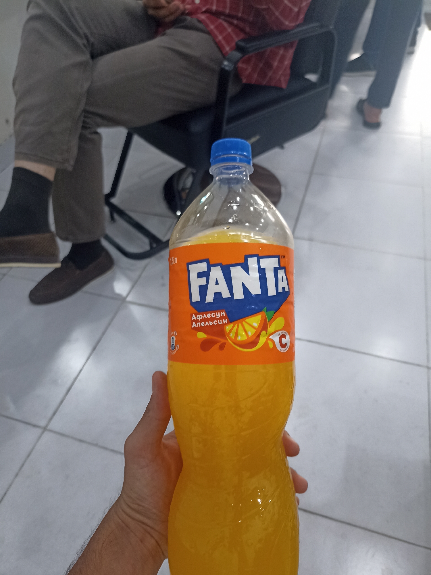 A person's hand is holding a clear plastic bottle of Fanta orange soda. The bottle is approximately two-thirds full of orange liquid and has an orange label with blue lettering. The label prominently displays the word "FANTA" in white letters. The background of the image shows a person seated in a black office chair with their legs crossed, wearing grey pants and a red and white checkered shirt. The floor is tiled with white and grey squares. The scene appears to be indoors, possibly in an office or waiting area, in the city of Dushanbe, Tajikistan. The lighting is bright, suggesting an indoor setting with artificial light.