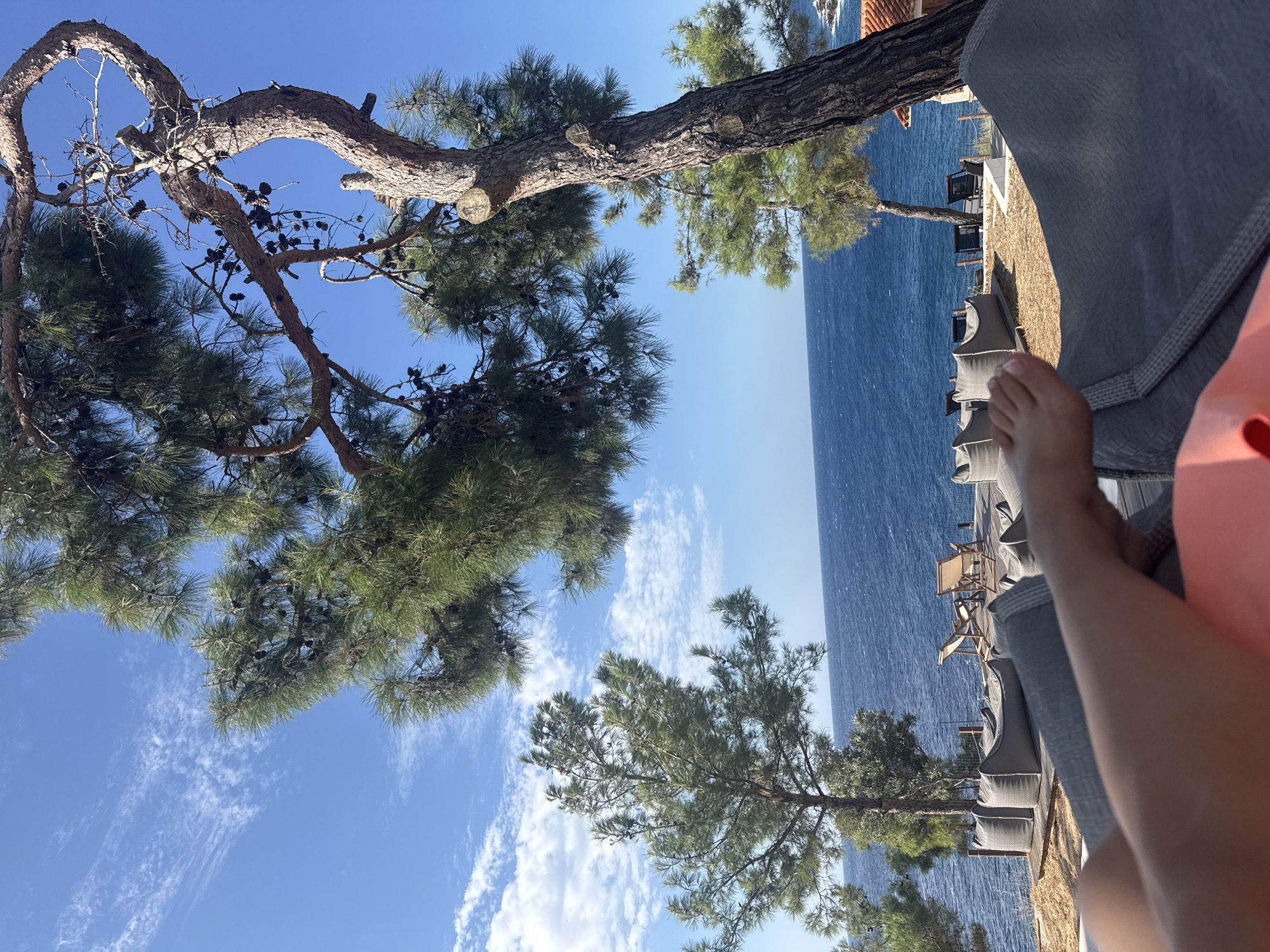 The image depicts a person's leg and foot resting on a grey beanbag chair in the foreground. Behind the chair, a wooden deck overlooks the deep blue sea under a bright blue sky dotted with white clouds. Several other beanbag chairs and lounge chairs are scattered across the deck. Lush green pine trees frame the scene, with one large pine tree on the right side of the image featuring prominent cones. The location is identified as Unknown, Greece. The overall impression is a serene, outdoor relaxation setting with a scenic ocean view.