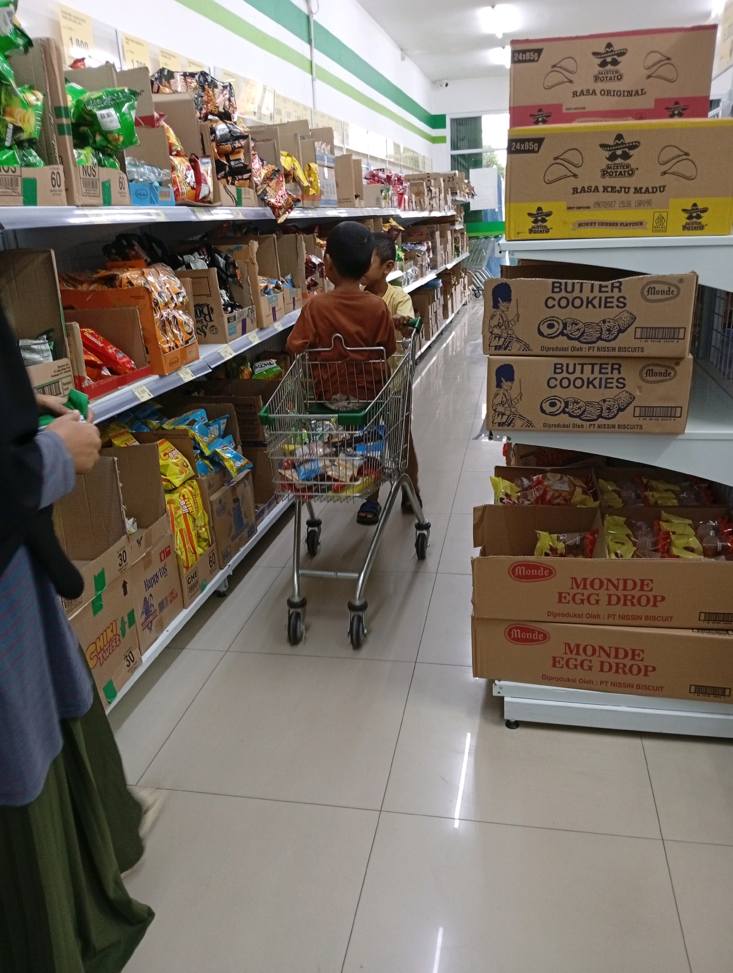 In Babakan, Indonesia, within a supermarket aisle, two young boys are depicted. The boy in the foreground, wearing a brown shirt, is pushing a shopping cart. The cart is filled with various packaged goods, including snacks and cookies. Another child, partially visible behind him, is also present. The aisle is lined with shelves stocked with boxes and bags of products. To the right of the shopping cart, stacked boxes of "Monde Egg Drop" cookies are visible. A person, likely an adult, is partially visible on the left side of the frame, wearing a blue and black striped shirt and a green skirt, holding a green item. The floor is tiled and reflects overhead lighting.