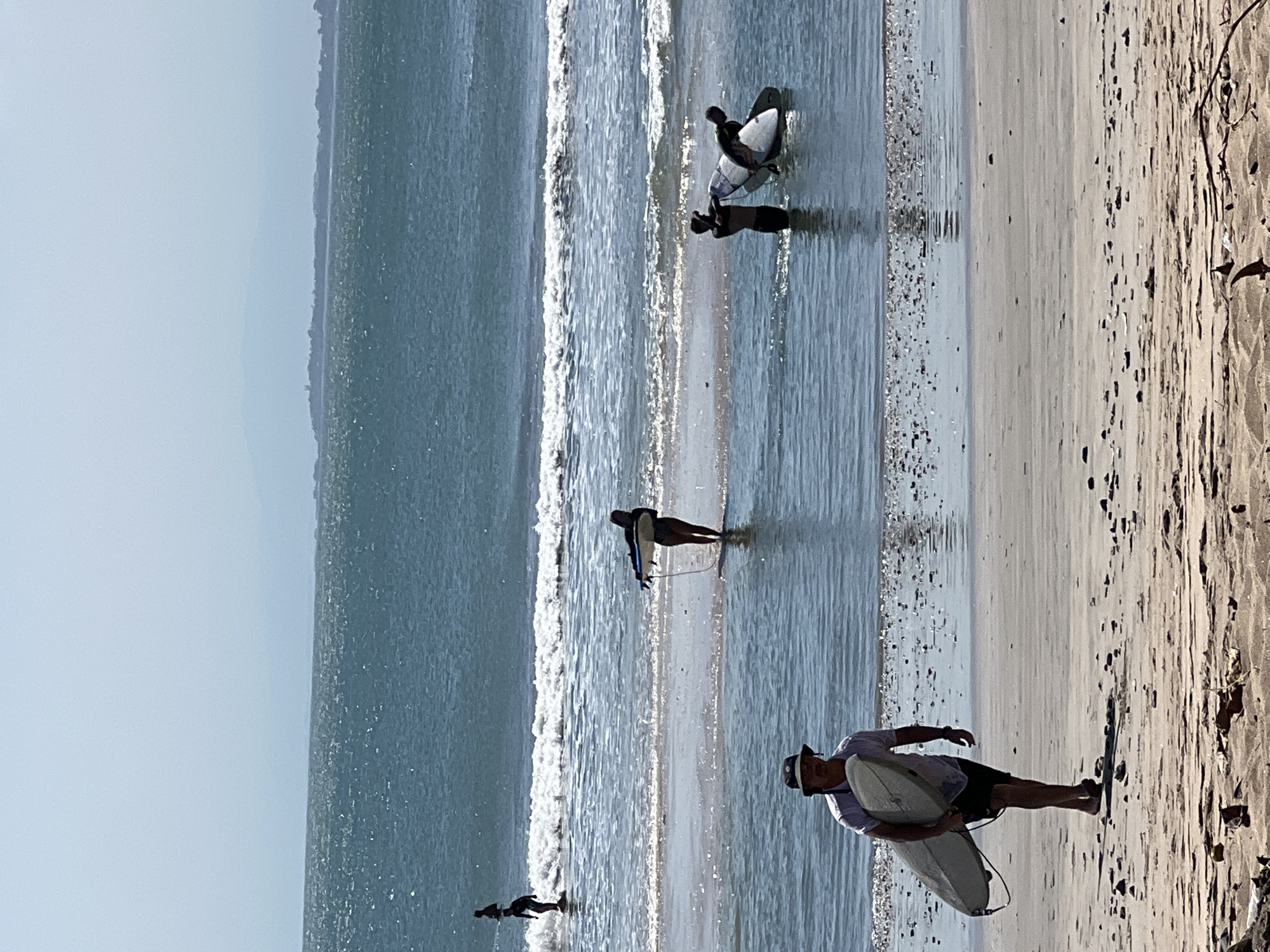 In Kuta, Indonesia, a beach scene unfolds under a hazy sky. In the foreground, a man wearing a bucket hat and a white t-shirt walks along the sandy shore, carrying a light-colored surfboard under his left arm. He is heading away from the water, his shadow stretching out behind him. The beach is strewn with small pebbles and debris.
Further into the water, several individuals are engaged with surfboards. Two people stand in the shallow water, one appearing to hand a surfboard to the other. A third person, further out, is standing upright on a surfboard in the gentle waves. In the mid-ground, a lone surfer stands in the water, holding a surfboard. In the distance, a break in the waves is visible, and the hazy horizon shows land formations. The ocean is a deep blue, with whitecaps on the waves.