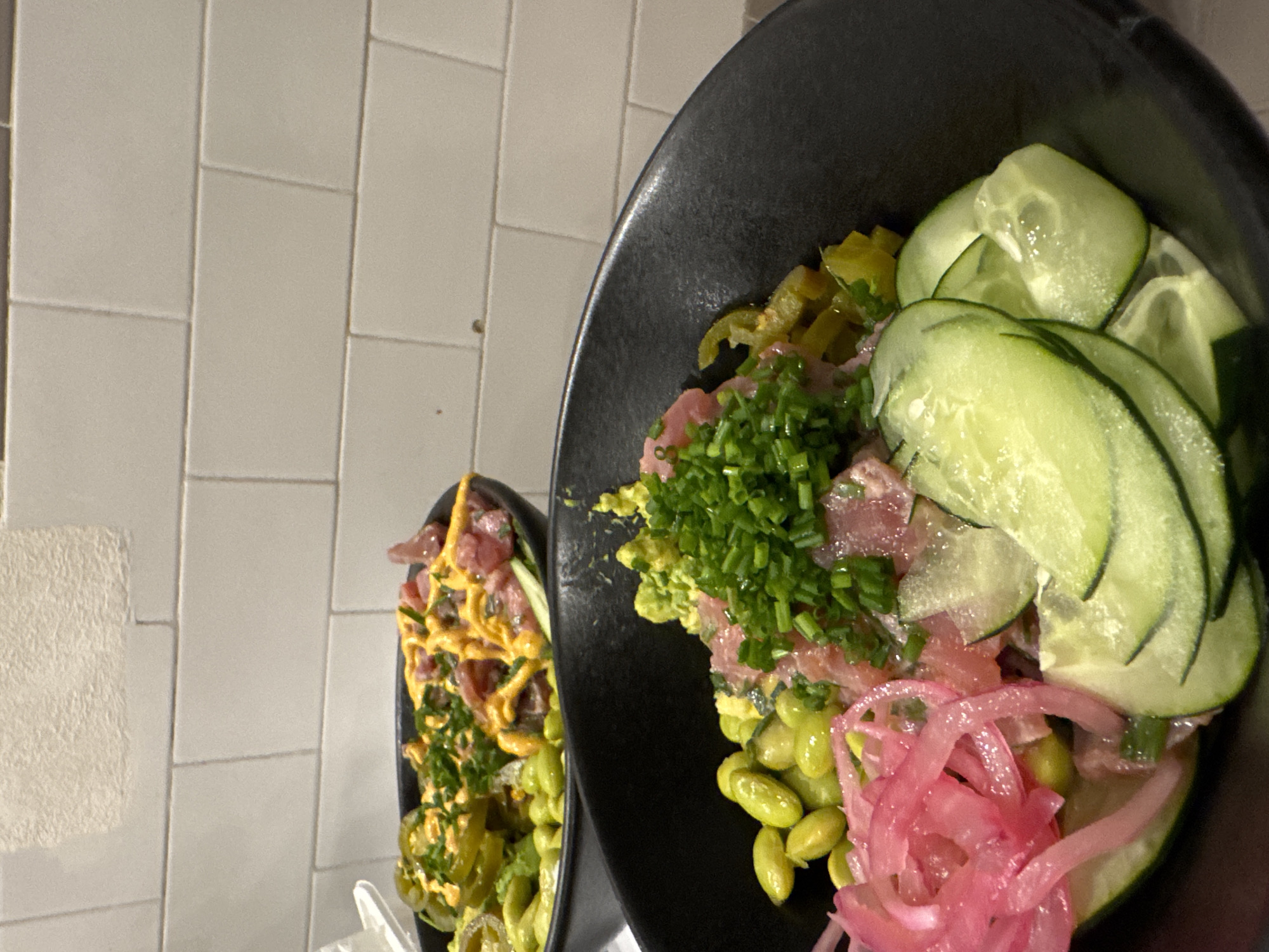 The image shows two bowls of what appears to be poke. The bowl on the right contains edamame, sliced red onion, cubed raw fish, chopped chives, sliced cucumbers, and a green purée. The bowl on the left contains edamame, sliced jalapeños, cubed raw fish, chives, and a yellow sauce. Both bowls are black and set on a table in front of a grey tiled wall. The location is likely a restaurant, potentially in Mexico City, Mexico.