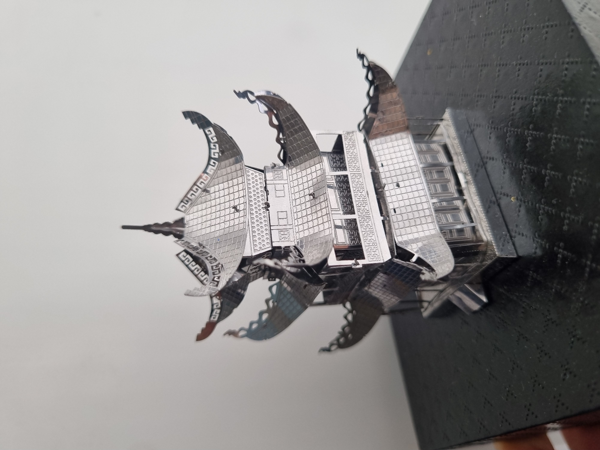 The image depicts a metal model of a building. It has a tower-like structure, and curved elements with Greek-like patterns. The model is primarily silver-colored. The building design shows windows and details which are partially open/empty. The model is fixed to a black square base. The background is mostly white on the left side.
There is no information to determine the location in the city of Chaikovsky, Russia.