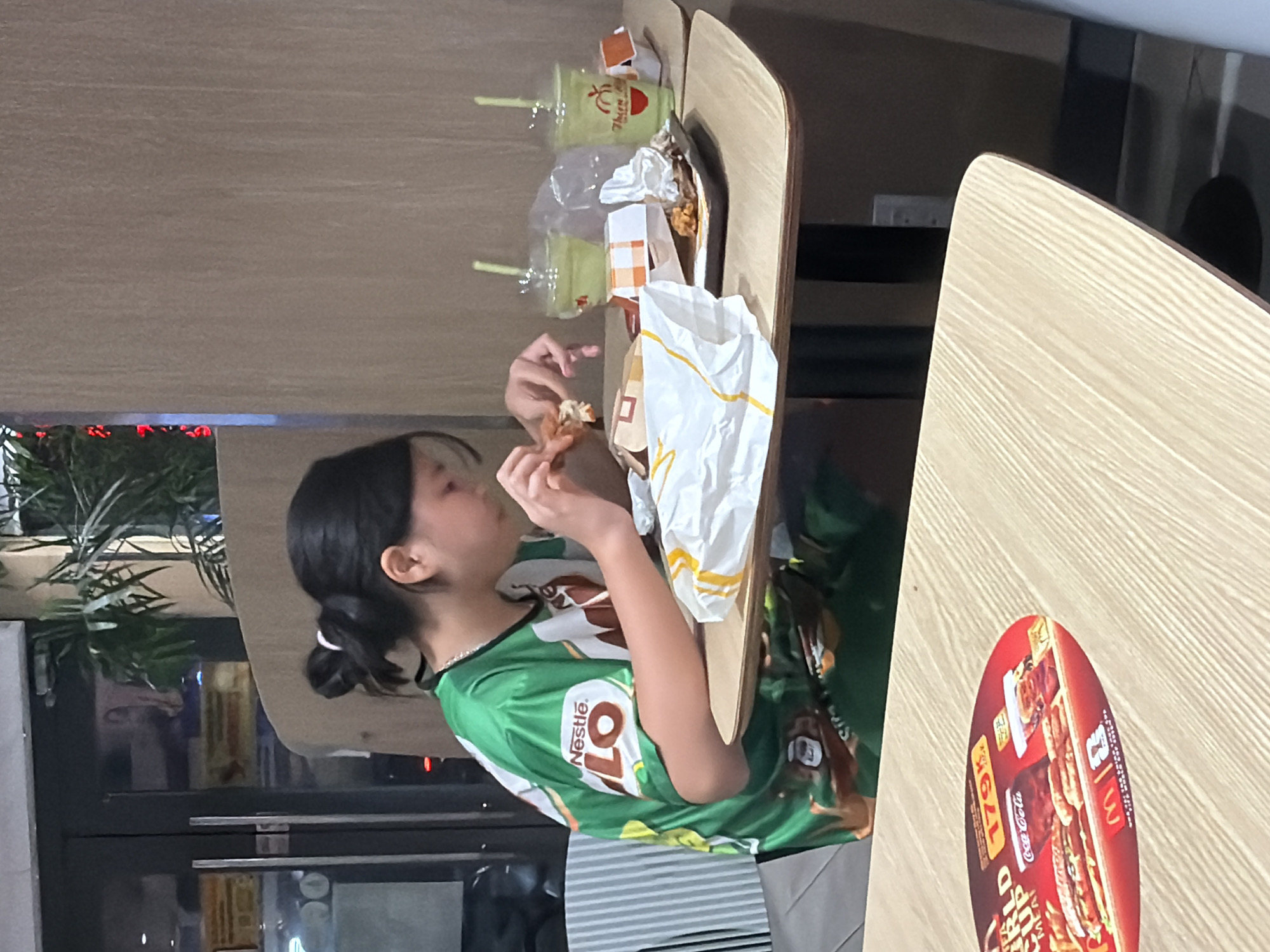 A young person, appearing to be female with dark hair pulled into a bun, sits at a light-colored wooden table in what appears to be a McDonald's restaurant. The individual is wearing a green shirt with white and brown accents featuring the "Nestle Milo" logo. They are actively eating, holding a piece of food in their hands.
On the table in front of them are several items: a white paper bag with orange and yellow stripes, a small orange and white checkered box, a tray with some fried chicken pieces and crumpled white paper, and two clear plastic cups with green liquid and straws. A large, circular red placemat with images of food and the Coca-Cola logo is visible on a table in the foreground, partially obscuring the lower part of the scene. The background shows a neutral-colored wall and a glimpse of plants and glass doors with reflections. The lighting suggests an indoor, possibly commercial setting. This scene is located in Nha Trang Ward, Vietnam.