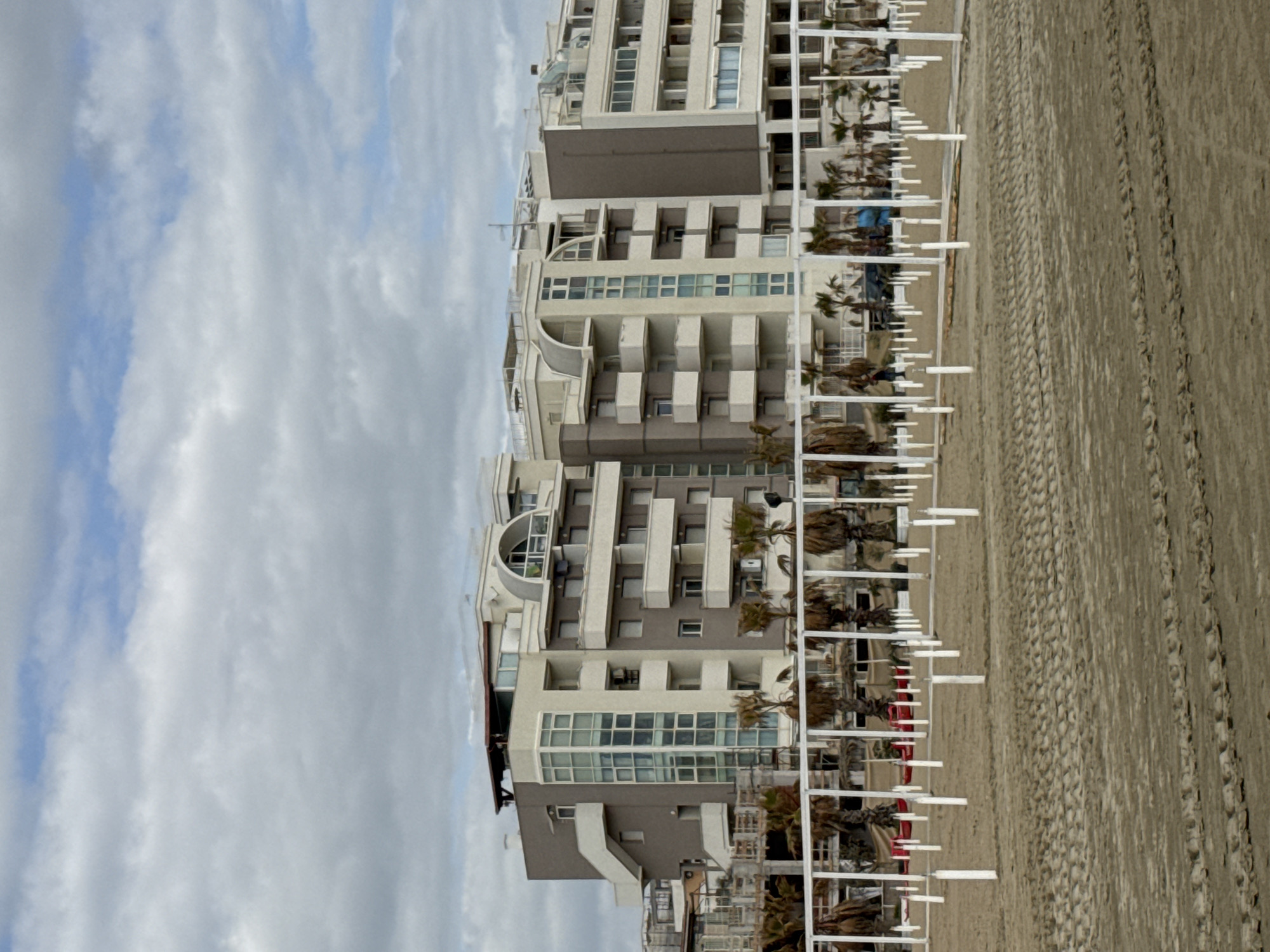 The image, when viewed in its correct orientation (rotated 90 degrees counter-clockwise), displays a coastal scene featuring multi-story buildings, a sandy beach, and a cloudy sky.
On the left side of the frame, several modern-style buildings, likely apartments or hotels, line the coast. These structures are primarily light-colored (white, beige, and light grey) with some darker grey accents. They feature numerous balconies, many with glass railings, and a variety of windows, some large and panoramic, others smaller. Architectural details include curved sections on some buildings. Palm trees are visible adjacent to the buildings, adding a tropical touch to the landscape.
To the right of the buildings lies a wide, expansive sandy beach. The sand is marked with numerous tracks and indentations, possibly from vehicles used for beach maintenance or from foot traffic. Along the edge of the beach, parallel to the buildings, runs a series of white, vertical posts, which appear to be part of a structured beach setup, possibly for umbrellas or private beach areas. Hints of red material are visible beneath some of these white structures.
The sky overhead is largely overcast with varying cloud formations, though patches of blue sky are visible through the clouds. The lighting indicates it is daytime. The absence of people and active use suggests the image might have been taken during an off-peak season or a quiet time of day. No discernible text is visible in the image. The scene is typical of a developed Mediterranean coastline, aligning with the provided location context of Nettuno, Italy.