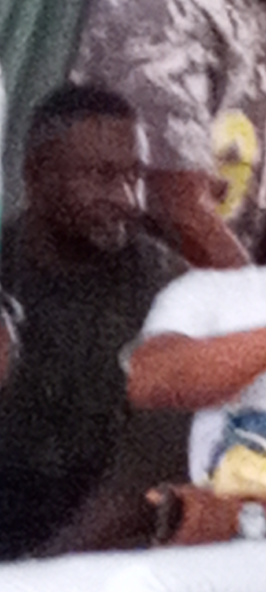 The image shows a person with dark skin wearing a dark-colored shirt. Their hand is raised to their face. Another person is partially visible, wearing a white shirt with a design on it. The background is blurred and indistinct.