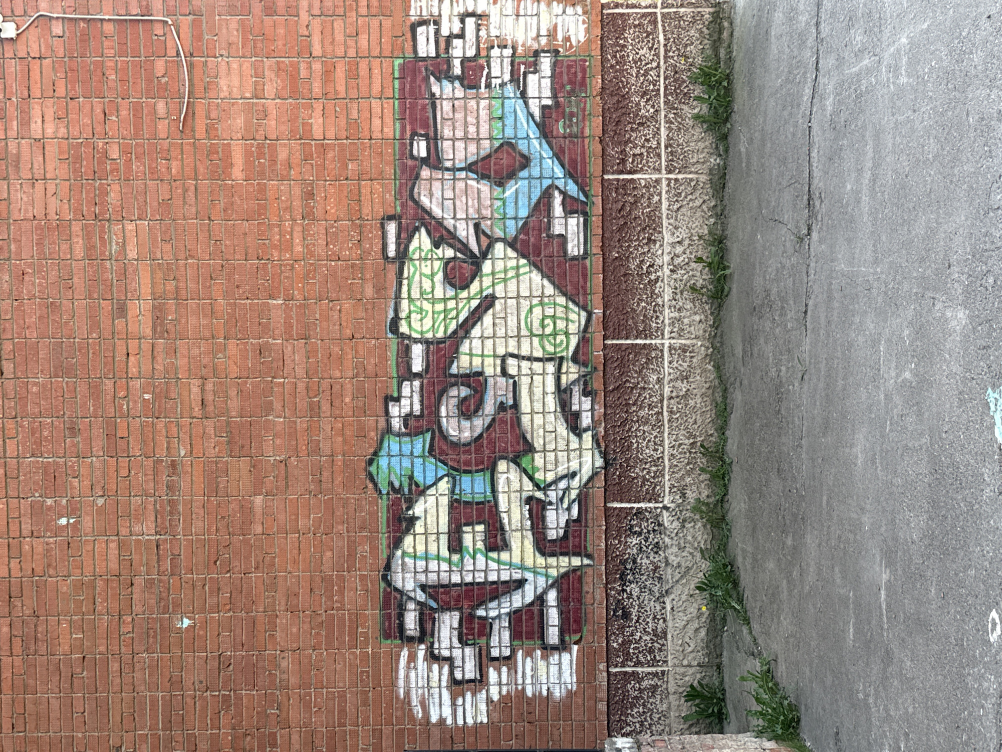 Viewed from above, the image depicts a section of a brick wall adorned with graffiti, adjacent to a pavement in Votkinsk, Russia. The brick wall is predominantly red with a section of patterned tiles along the right side. Graffiti, comprised of abstract shapes and colors including blue, green, and maroon, is painted on the brick wall section. The pavement, gray in color, abuts the patterned tiles, with vegetation growing in the crack between them. A white electrical cord runs along the upper left portion of the brick wall.
