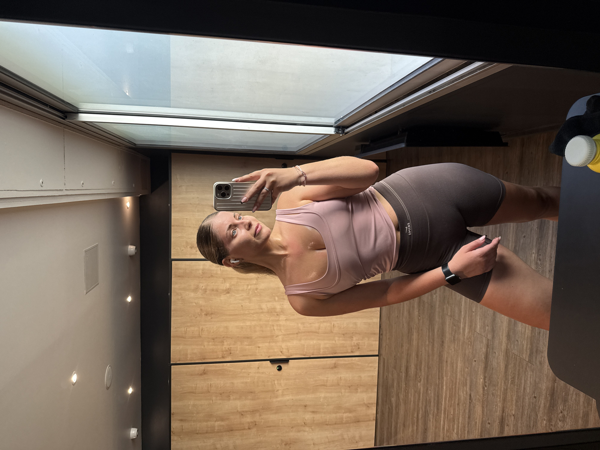 The image shows a woman taking a selfie in a gym locker room. She is wearing a pink sports bra and brown athletic shorts, with white wireless earbuds in her ears. She has a fitness watch on her left wrist. The locker room has wooden paneling on the walls and a light-colored ceiling with a skylight. There are spotlights on the wall and a vent. The woman appears to be sweaty, suggesting she has just finished a workout. The image is taken in a vertical orientation. The setting implies a modern gym, possibly in Paris, France, given the location context.
