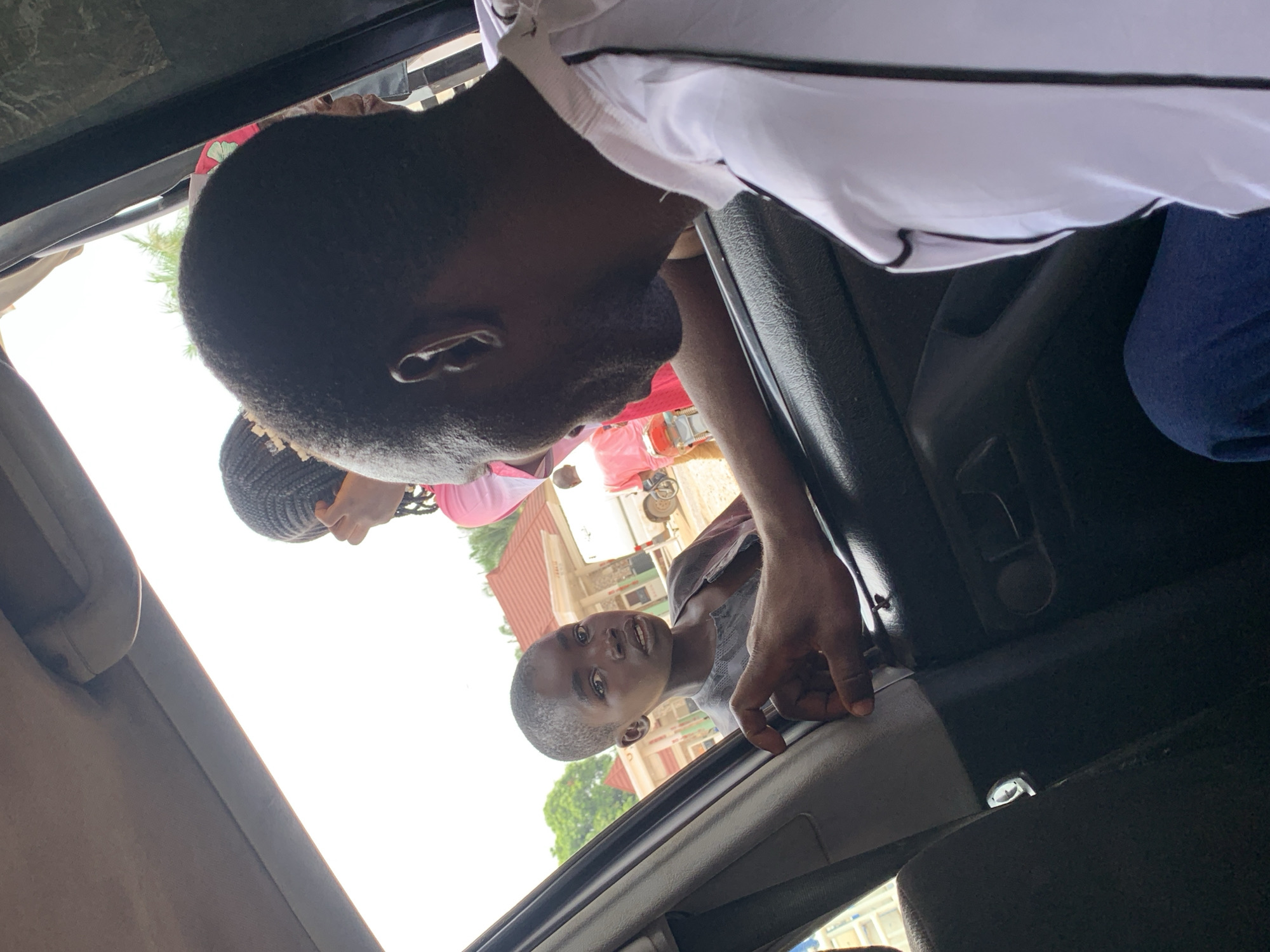 A low-angle shot taken from inside a car, with the camera positioned slightly above the interior. A man leans into the car's interior, wearing a white shirt, with his head positioned at the top center of the frame. Below and to the left, another man looks up towards the car from the outside, with a residential building and a parked vehicle in the background. A woman with braided hair is visible partially to the left of the first man, suggesting she is standing near the car. The photo was taken in Akwanga, Nigeria.
