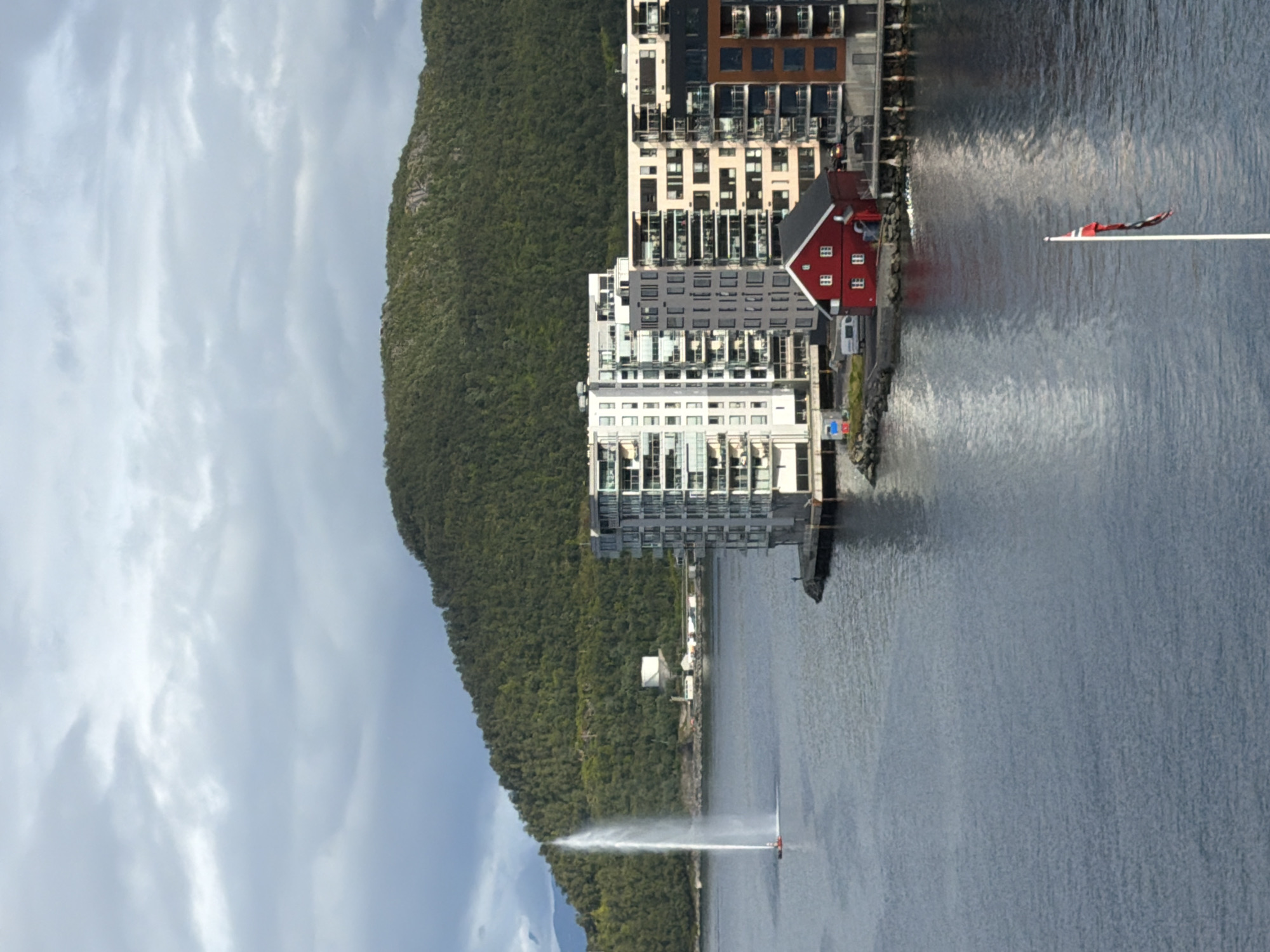 The image depicts a body of water with a tall water fountain on the left, likely a boat or ship emitting the spray. In the background, a lush green, tree-covered hill rises steeply. On the right side of the water, a row of modern, multi-story buildings with balconies forms a waterfront cityscape. A small, red traditional Norwegian house with white-framed windows is situated in front of these buildings. Further to the right, a section of a building with a brown facade is visible. In the foreground, a flagpole with a Norwegian flag is planted in the water. The sky is overcast with dramatic clouds. The overall scene suggests a port or coastal area in a city, potentially Harstad, Norway, given the presence of the Norwegian flag and traditional architecture. There is a single person visible standing on the dock in front of the buildings. A white van is parked near the red house.