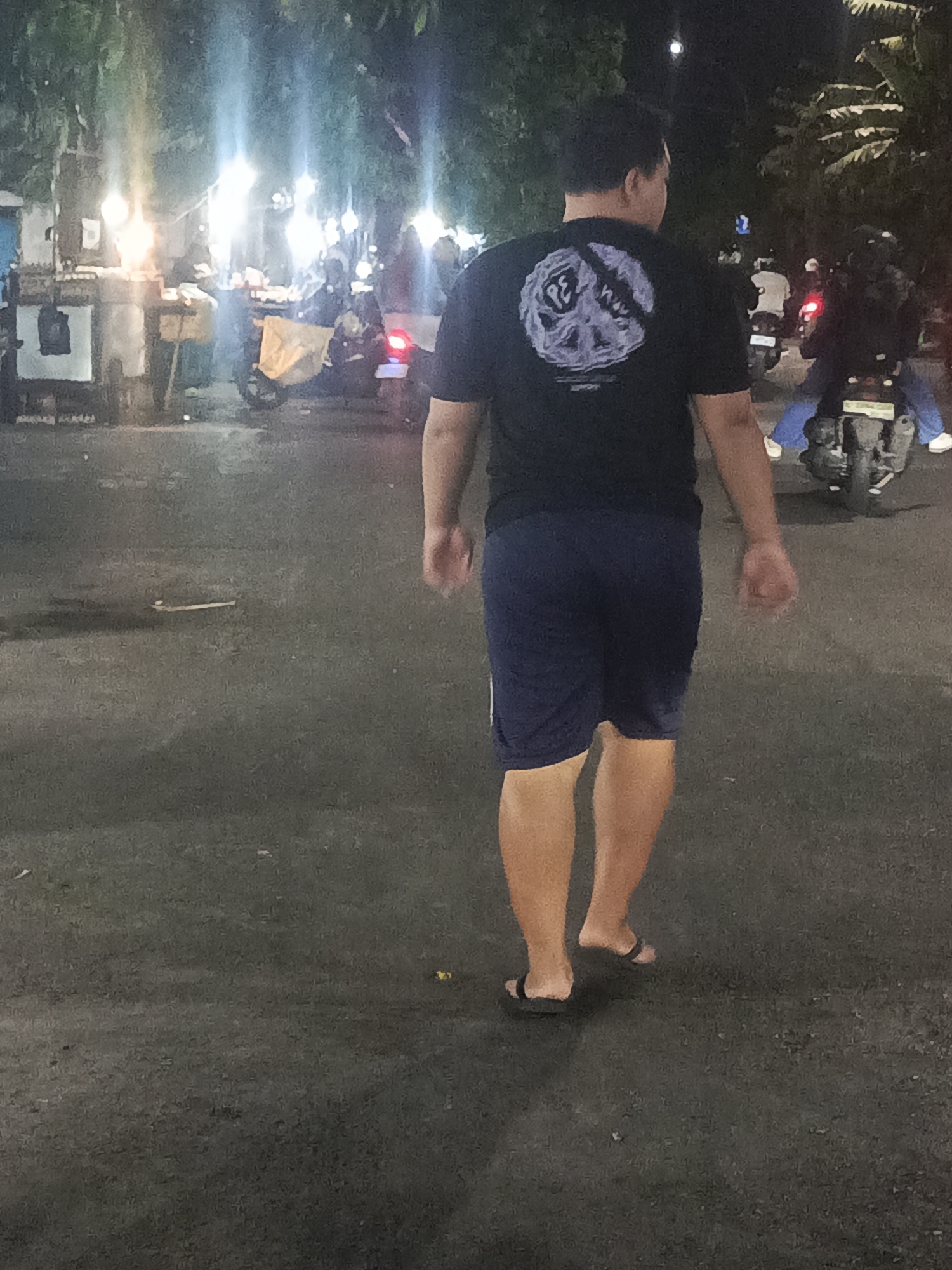 A full shot shows a man walking away from the camera on a dark asphalt road. The man has short, dark hair and is wearing a black t-shirt with a graphic design on the back and navy blue shorts. He is wearing black flip-flops. The road appears to be a public area, with a few other people on motorcycles in the background. The scene is lit by artificial lights, suggesting it is nighttime or dusk. The background includes some trees and what appears to be food stalls or market stalls, illuminated by bright lights. The location is the street in front of a market in the Rungkut district of Surabaya, Indonesia.