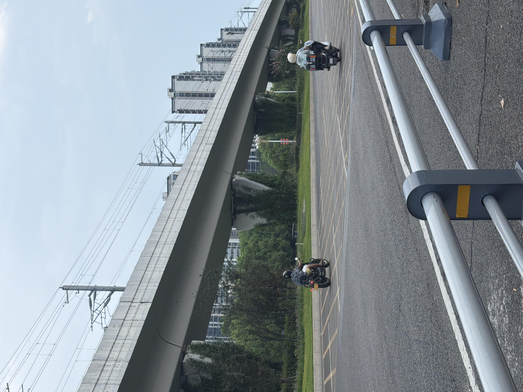 The image shows a scene in the city of Jiangjin, China, featuring a viaduct and surrounding urban landscape. In the foreground, a two-wheeled motor vehicle with two occupants is visible on a paved road. The vehicle has a black box on the rear. The driver and passenger are wearing helmets. Further in the background, another motor vehicle with a single rider is also on the road. This vehicle also appears to be a motorcycle. Both vehicles are in motion, moving away from the viewer. The viaduct, a prominent concrete structure, spans across the scene, supported by columns. Overhead power lines are visible on the viaduct. Behind the viaduct, a row of tall, modern residential buildings with a light-colored facade and dark windows can be seen. Green trees and vegetation are present between the road and the buildings, with some ivy growing on the viaduct's supporting columns. The sky is overcast. In the immediate foreground, a grey metal guardrail with yellow accents is present. The road surface is asphalt with yellow lane markings. The overall impression is an urban environment with elevated infrastructure and residential development.