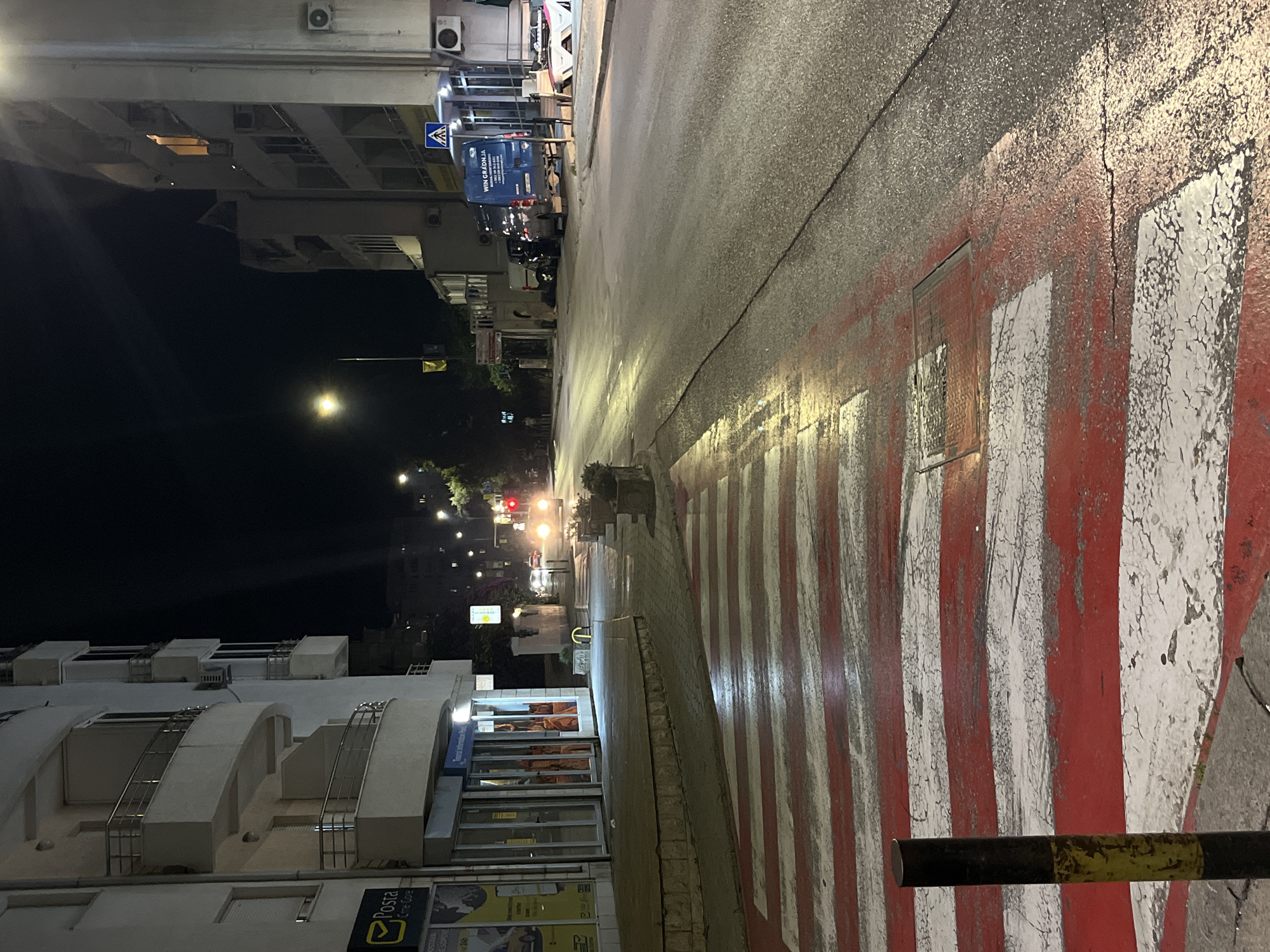 The image captures a street scene at night in Budva, Montenegro. A crosswalk with red and white stripes dominates the foreground. The street is wet, reflecting the overhead lights. On the left, a building with balconies and commercial storefronts, including a "Posta" sign and "Tourist Information Point," is visible. Across the street, more buildings are present, with streetlights illuminating the roadway. Several vehicles, including a blue van with "WIN GRADNJA" written on it, are parked along the curb. In the distance, headlights of approaching cars can be seen. The overall environment suggests a quiet evening in an urban setting.
