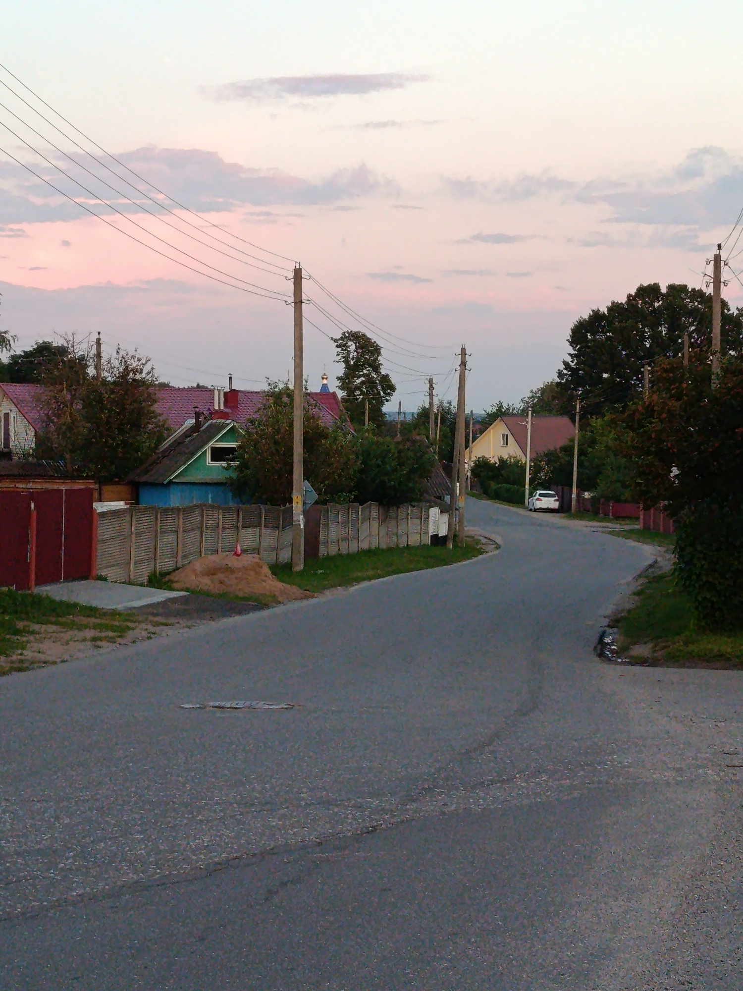 The image depicts a rural road in Смілавіцкі сельскі Савет, Belarus, during the twilight hours. The road is asphalt and curves to the right, flanked by vegetation and residential structures. On the left, a red metal gate is visible, leading to a property with a wooden fence. Further down the road, several houses with reddish-brown roofs and some with green accents are nestled amongst trees. Power lines are strung along poles that line the road. A white car is parked on the side of the road near a yellow house. The sky is a soft gradient of pink and pale blue, indicating the end of daylight. The overall impression is one of quiet, rural life at dusk.
