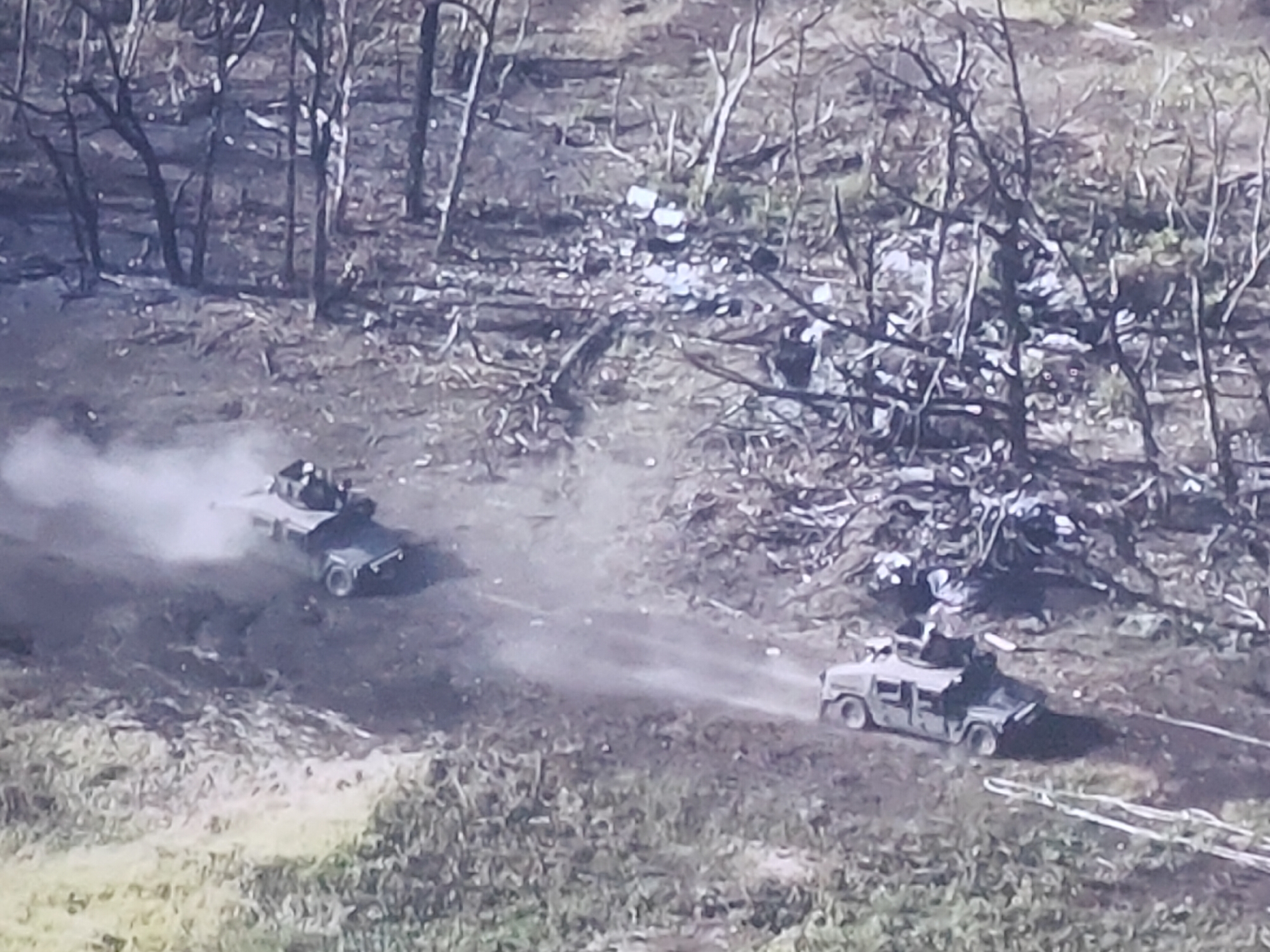 The aerial image shows two military vehicles, possibly Humvees, driving on a dirt road through a wooded area. The vehicle on the left is kicking up dust. The ground is covered with debris, including what appears to be broken branches and possibly some discarded items. The trees are bare, suggesting a cold season. There are no discernible buildings or urban features visible, making it impossible to confirm a location in Molenbeek-Saint-Jean - Sint-Jans-Molenbeek, Belgium, based solely on this image.