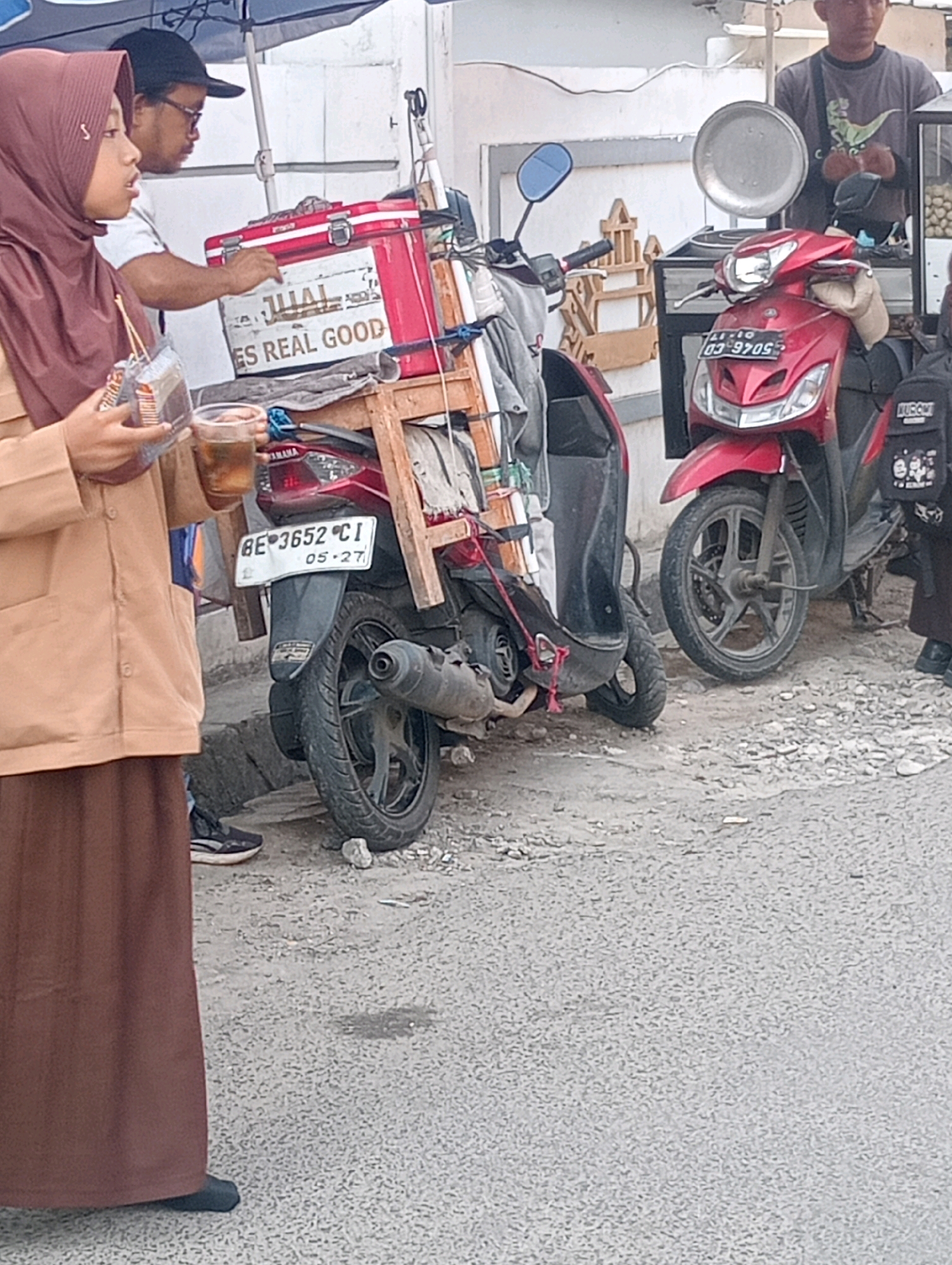 In Bandar Lampung, Indonesia, a young woman wearing a brown hijab and a tan jacket stands near a motorcycle with a red cooler box attached. She holds a clear plastic cup containing a dark liquid and a packet of food. A man wearing a black cap and glasses stands behind her, also near the cooler. A red scooter is parked to the right of the woman's motorcycle, with a metal tray and food containers visible on its handlebars. The scene appears to be a street-side food stall.