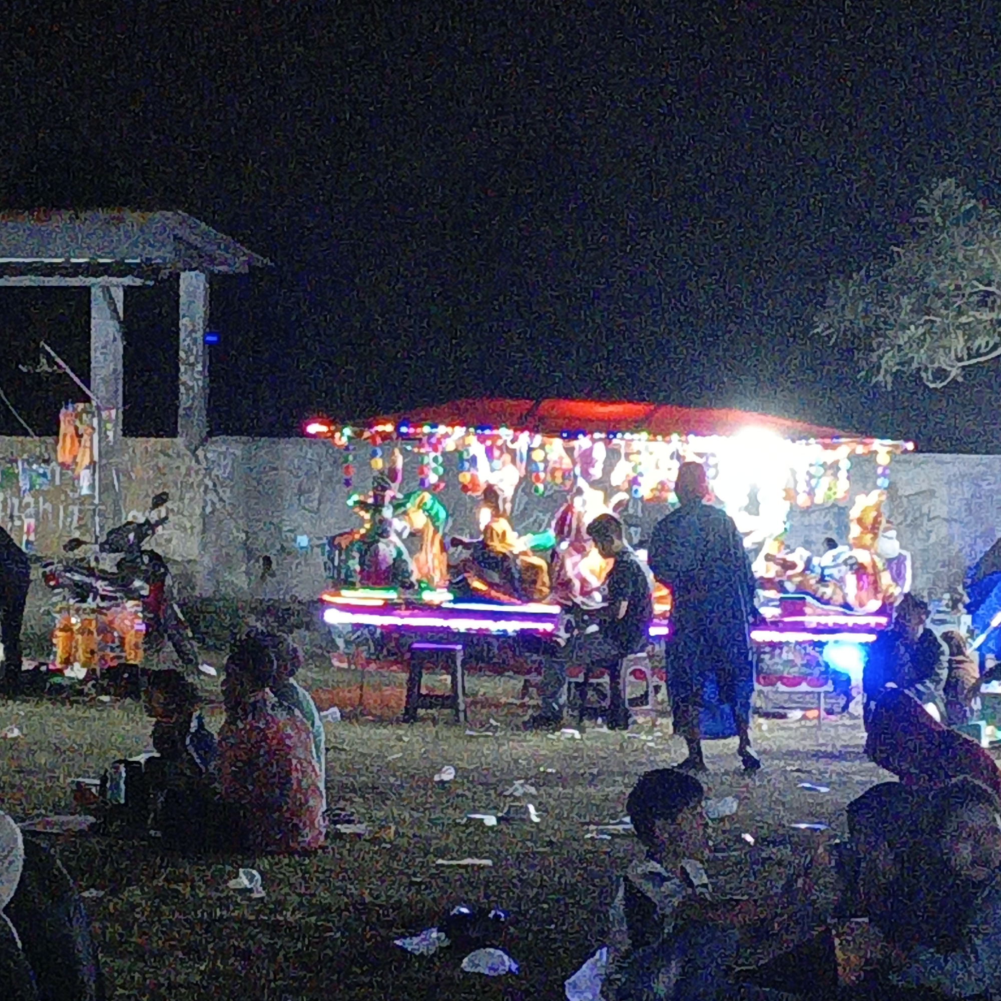 In Mangli, Indonesia, at night, an outdoor scene depicts a gathering around a brightly lit amusement ride. The ride, adorned with colorful lights, appears to be a carousel with various figures. Several people are visible, some seated on the ride, others standing and observing. The ground is covered in grass and littered with debris, including paper cups. A motorcycle is parked to the left of the frame. The background shows a stone wall and a structure with a roof, suggesting an open-air event space or park. The overall atmosphere is festive, illuminated by the artificial lights of the amusement ride.