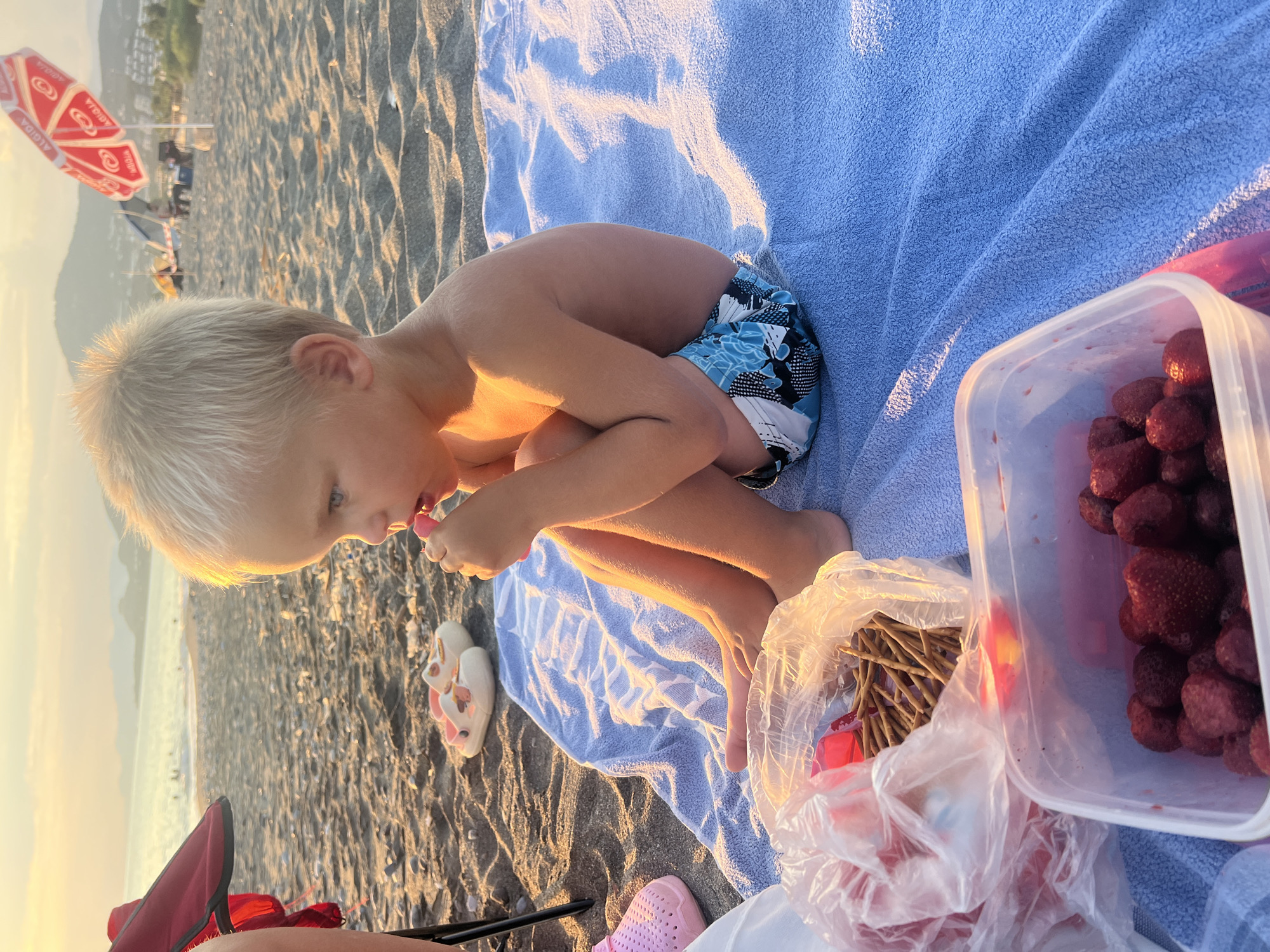 A young child with blonde hair is sitting on a blue towel on a sandy beach. The child is wearing blue patterned swim trunks and is holding a pink object to their mouth, possibly eating. To the right of the child is a clear plastic container filled with strawberries. In front of the child, also on the towel, is a plastic bag containing thin breadsticks. In the background, the sea is visible with waves, and further beyond, hills can be seen. Several beach umbrellas, one with red and white markings and text that appears to say "ALGIDA", are set up on the sand. The lighting suggests it is late afternoon or early evening, with the sun casting a warm glow on the scene. The overall mood appears relaxed and casual, indicative of a family outing at the beach. The location context provided is Alanya, Turkey.