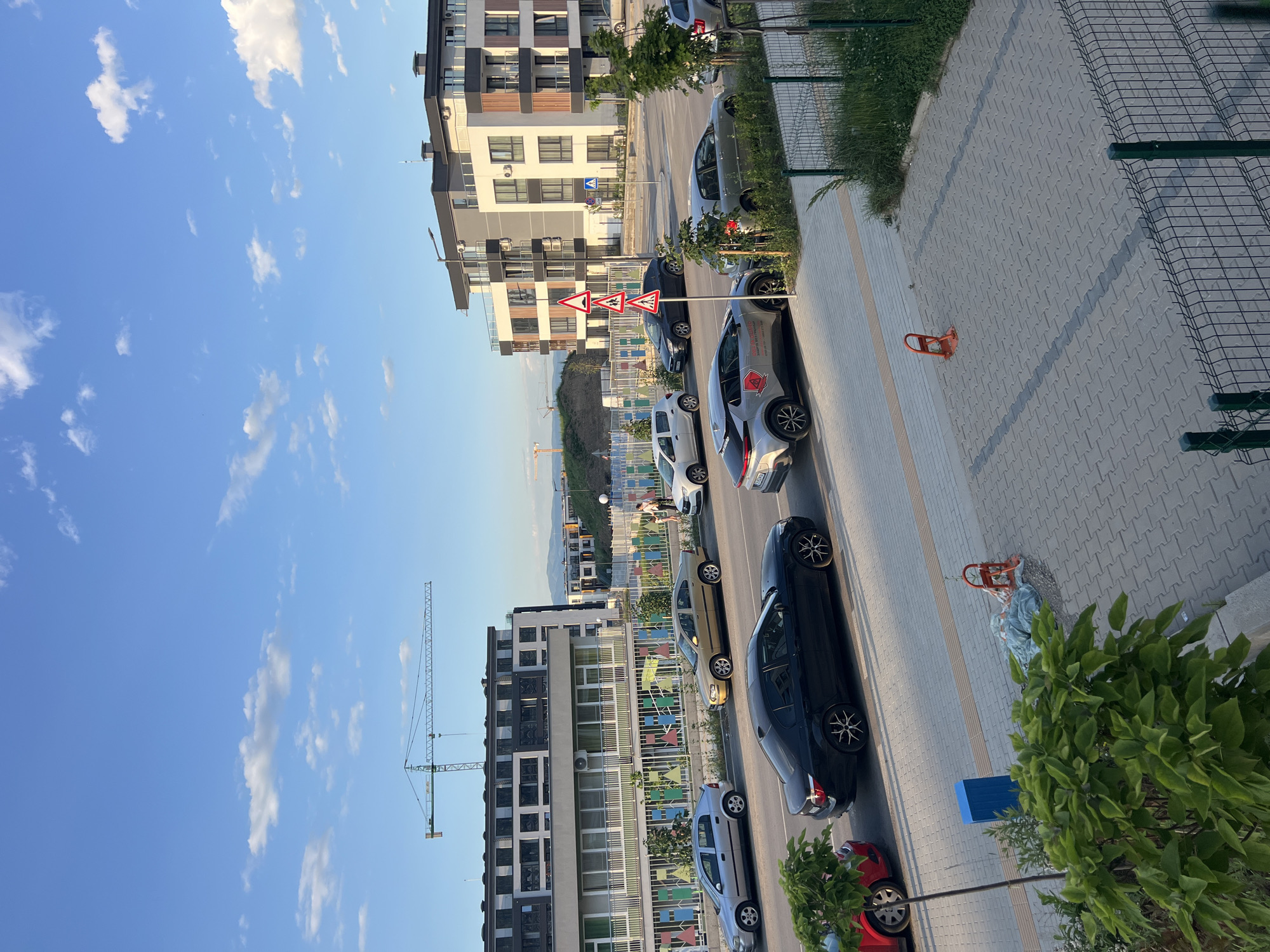 In an outdoor urban scene under a partly cloudy sky, a street is lined with parked cars and buildings. A green construction crane is visible in the background, along with other construction equipment and mounds of earth. In the foreground, several cars are parked along the street, including a silver hatchback, a gold sedan, a black sedan, and a silver crossover. A red car is partially visible on the left. Buildings with modern architecture, featuring balconies and large windows, are situated behind the street. A sidewalk runs alongside the street, with some greenery and trees planted along it. Traffic signs indicating speed bumps and pedestrian crossings are posted. There are a few people walking on the sidewalk in the distance. The ground is paved with interlocking stones and concrete.