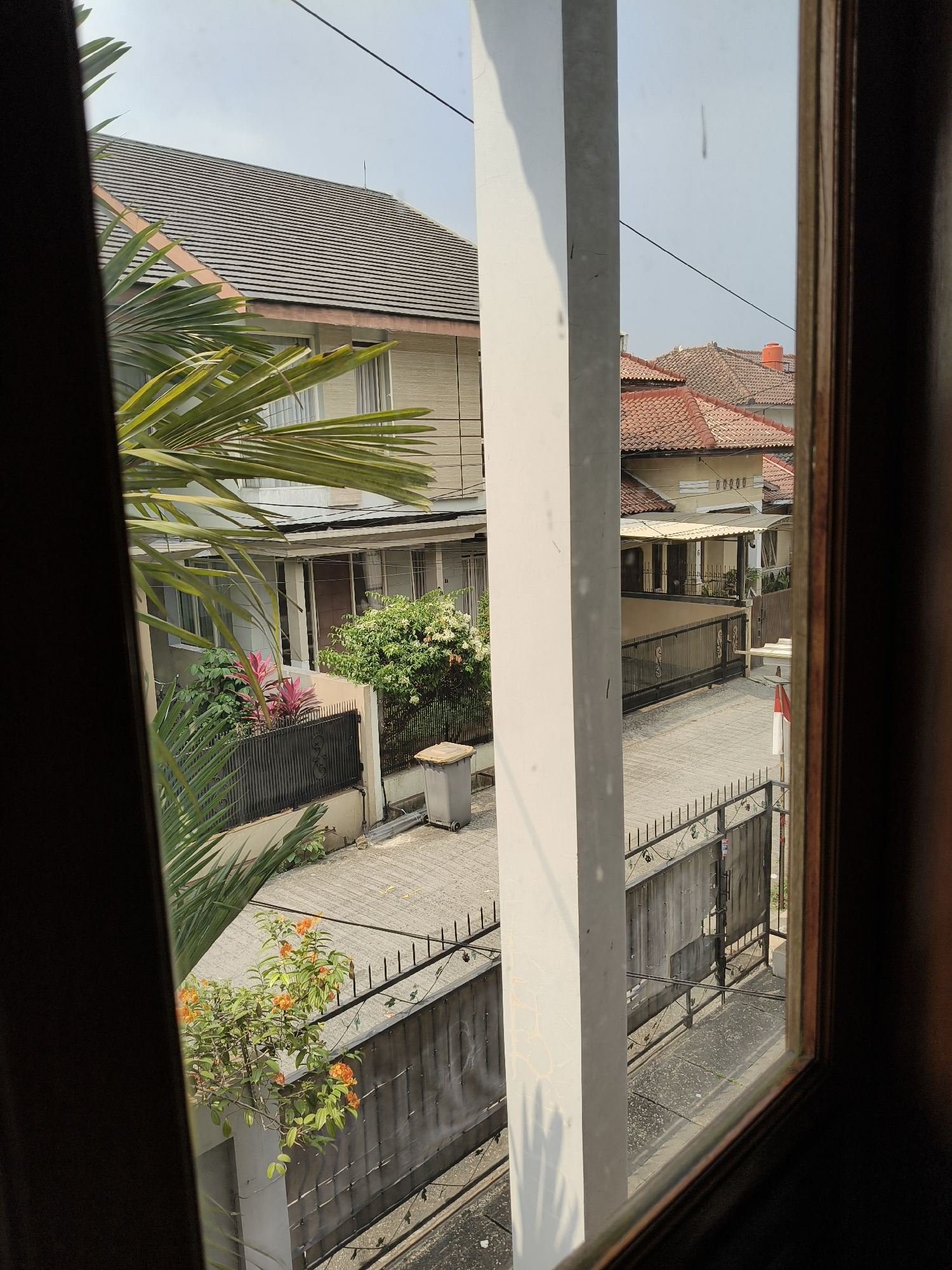 This image depicts a residential street view from an elevated perspective, possibly from a window or balcony. The scene is dominated by houses with tiled roofs, characteristic of urban or suburban Indonesian architecture, specifically in Bandung City. In the foreground, a large palm frond partially obscures the view from the left, while a white pillar runs vertically in the center.
The houses visible have multiple stories and are built close to each other. Some feature balconies and large windows. The street itself is paved with interlocking concrete blocks. A black metal fence runs along the property line of one house, with decorative elements. A grey trash bin is positioned near this fence. Beyond this, another fence, also made of metal, encloses a driveway or yard. In the distance, more houses with red tiled roofs are visible, suggesting a densely populated residential area. The sky is overcast, suggesting a hazy or cloudy day. There are no people or animals visible in the frame. The overall impression is a typical residential street scene in a tropical urban environment.