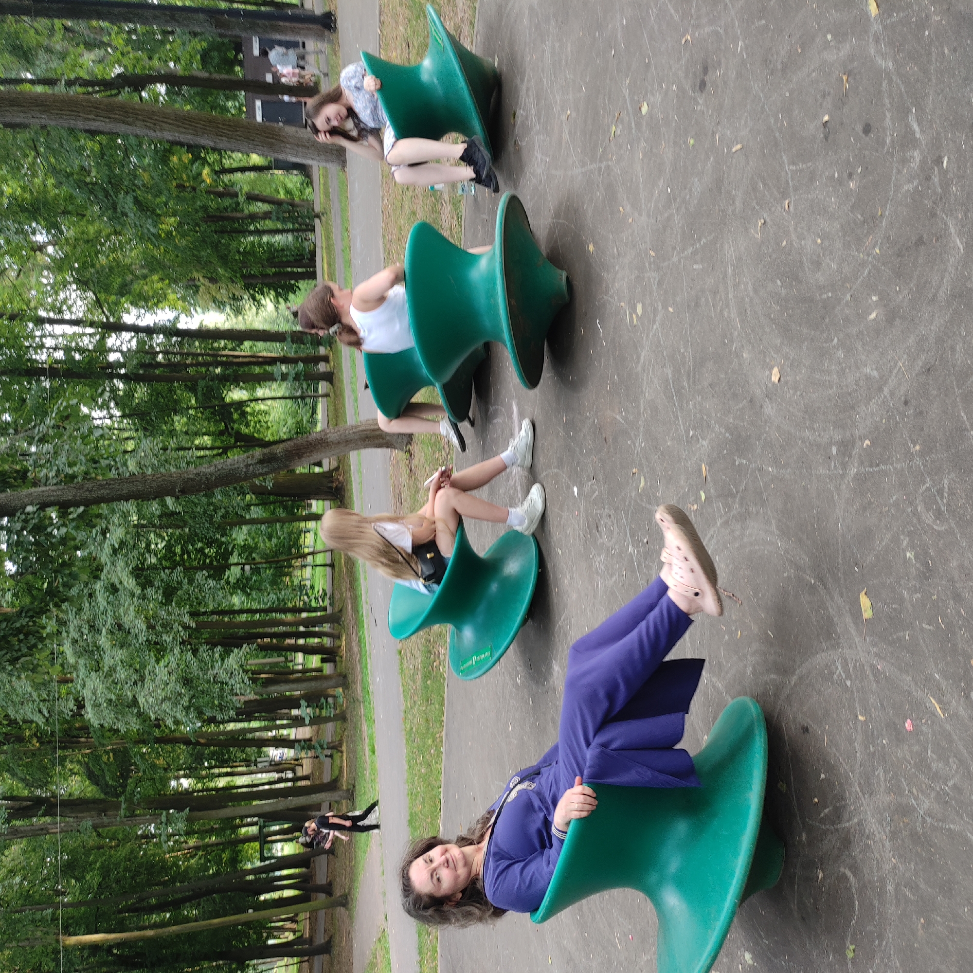 In a park setting with a backdrop of tall trees and a paved ground surface, four individuals are seated in unique, oversized green spinning chairs. A woman in a purple dress is in the foreground, seated and looking at the camera. To her right, a young girl with blonde hair sits in another chair, holding a white object. Further back and to the right, another young woman in a white top is in a spinning chair, touching her hair. The fourth chair is occupied by a young woman in a patterned dress, who is also looking to her right. In the background, a couple walks on a path, and other park-goers are visible in the distance near buildings. The scene appears to be set in a public park with distinct, modern playground equipment.