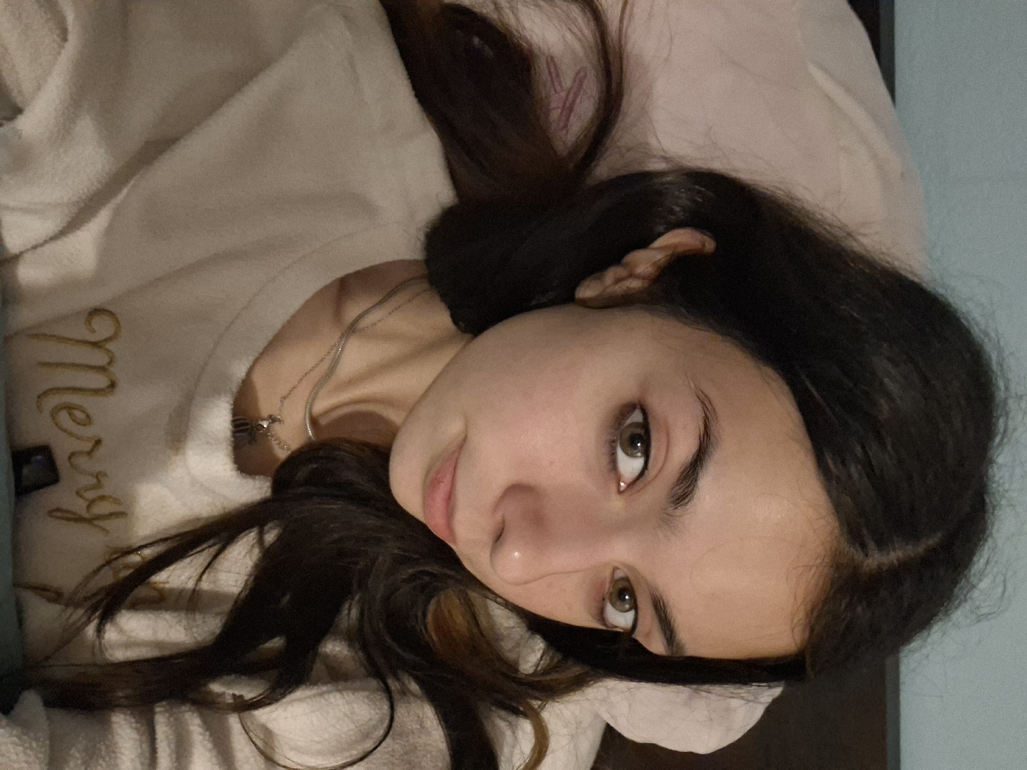 A young woman lies on a bed in a room with light blue walls. She is positioned on her back, head resting on a pillow. She has dark brown hair and brown eyes. She is wearing a light-colored, possibly white, long-sleeved shirt with gold lettering. A silver necklace is visible on her chest. The room's environment includes a bed, a pillow, a wall, and the clothing she is wearing. There is no indication of the image's location being Sofia, Bulgaria.