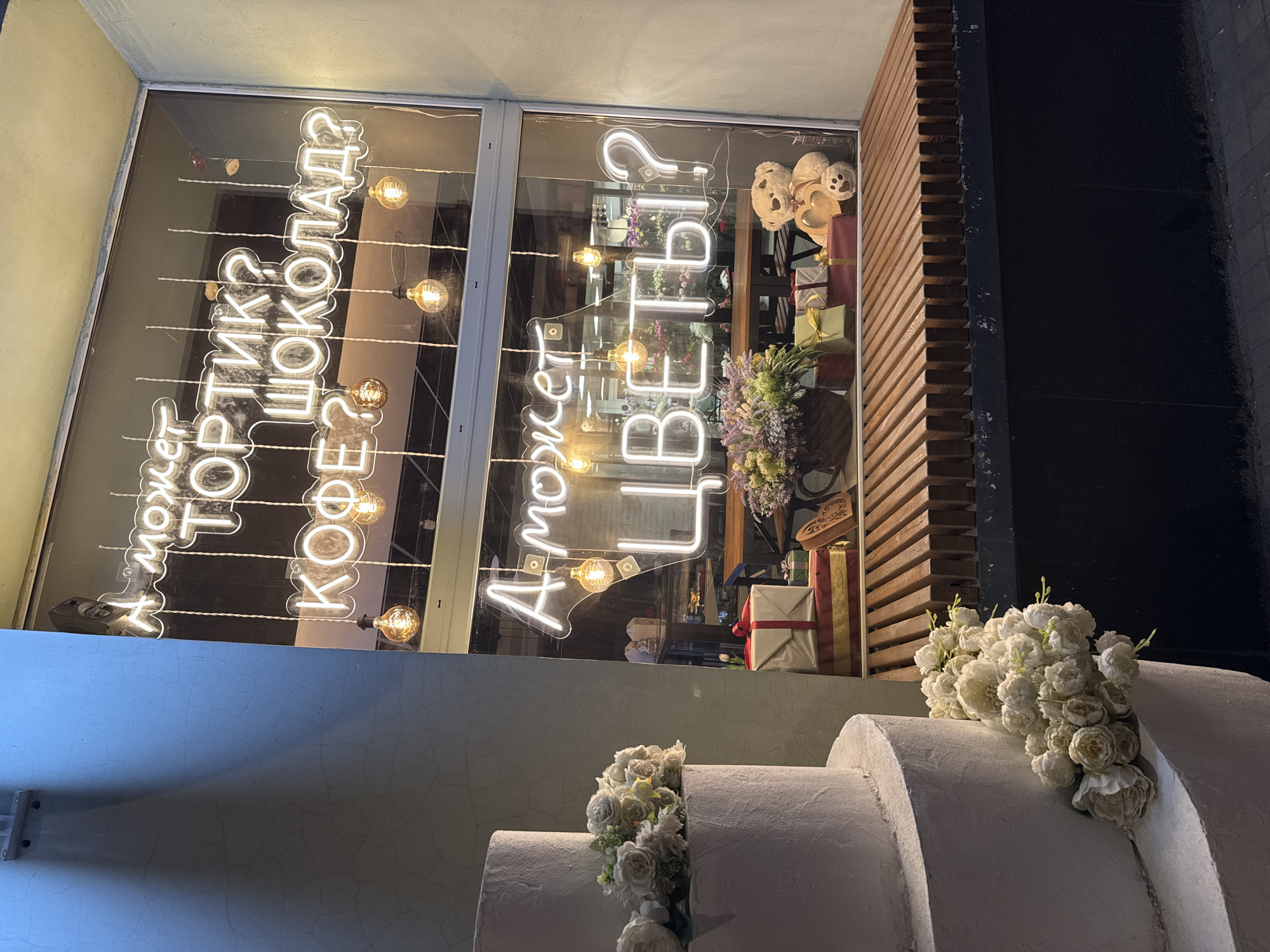 A nighttime street scene showing a shop window displaying neon signs and a tiered decorative cake. The neon signs, written in Cyrillic script, are illuminated in a warm white light and appear to be strung with vintage-style light bulbs. The sign on the left reads "А может тортик шоколад? кофе?" (Perhaps a chocolate cake? Coffee?). The sign on the right reads "А может цветы?" (Perhaps flowers?). The window display includes wrapped gifts, stuffed animals, and a bouquet of flowers. To the lower right, a tiered white decorative cake is adorned with white roses. The shop's exterior includes wooden slats on the right and a smooth, painted surface below the window. The location is unknown.