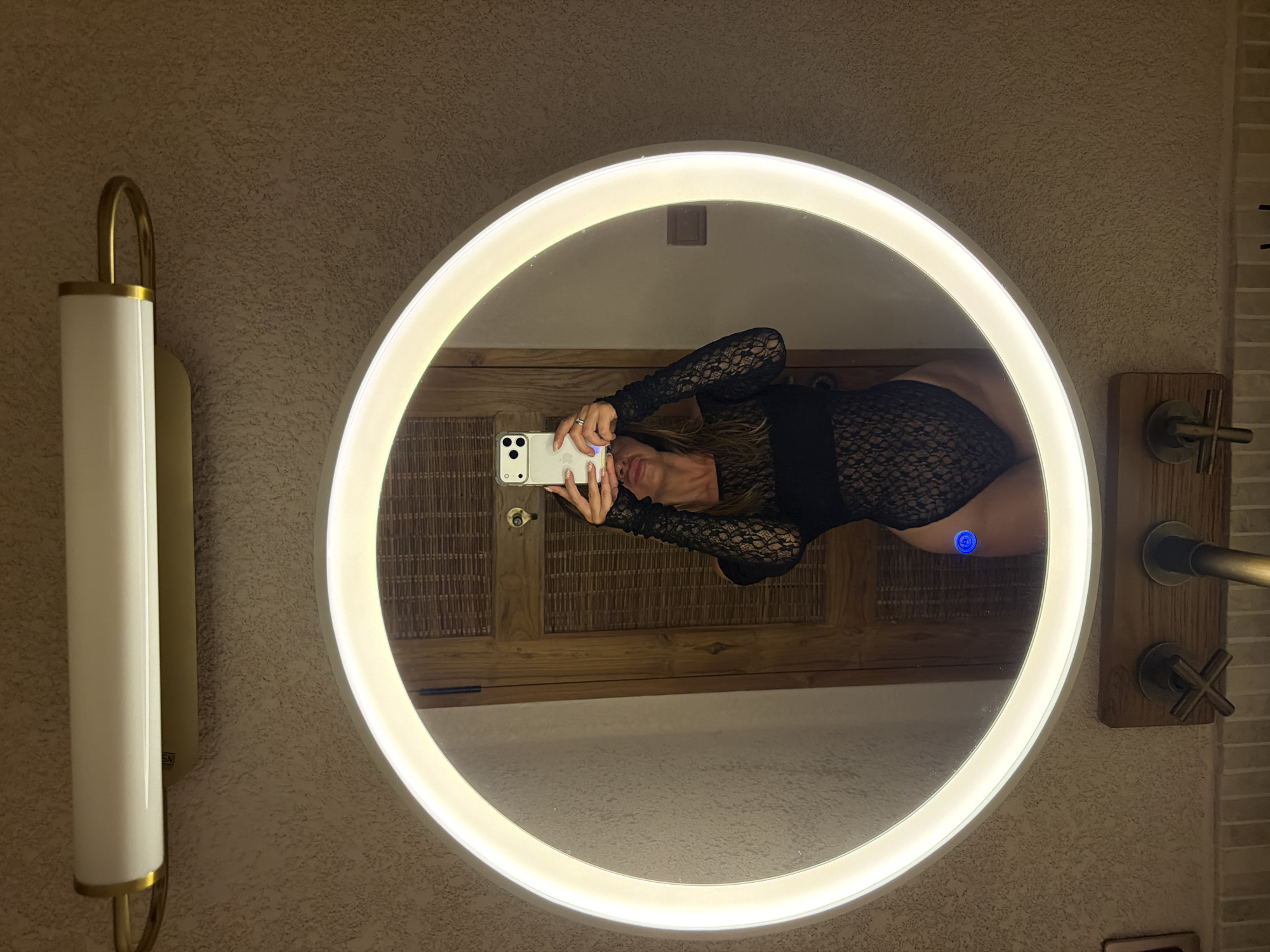 In a bathroom in Mengwi, Indonesia, a woman is taking a mirror selfie. She is wearing a black lace bodysuit and holding a smartphone in her right hand to capture her reflection. The bathroom features a large, circular mirror with a bright LED light ring, and a horizontal light fixture with a gold accent above the mirror. The wall has a textured finish, and below the mirror, a wooden fixture with faucets is visible. The background reflected in the mirror shows a wooden door.