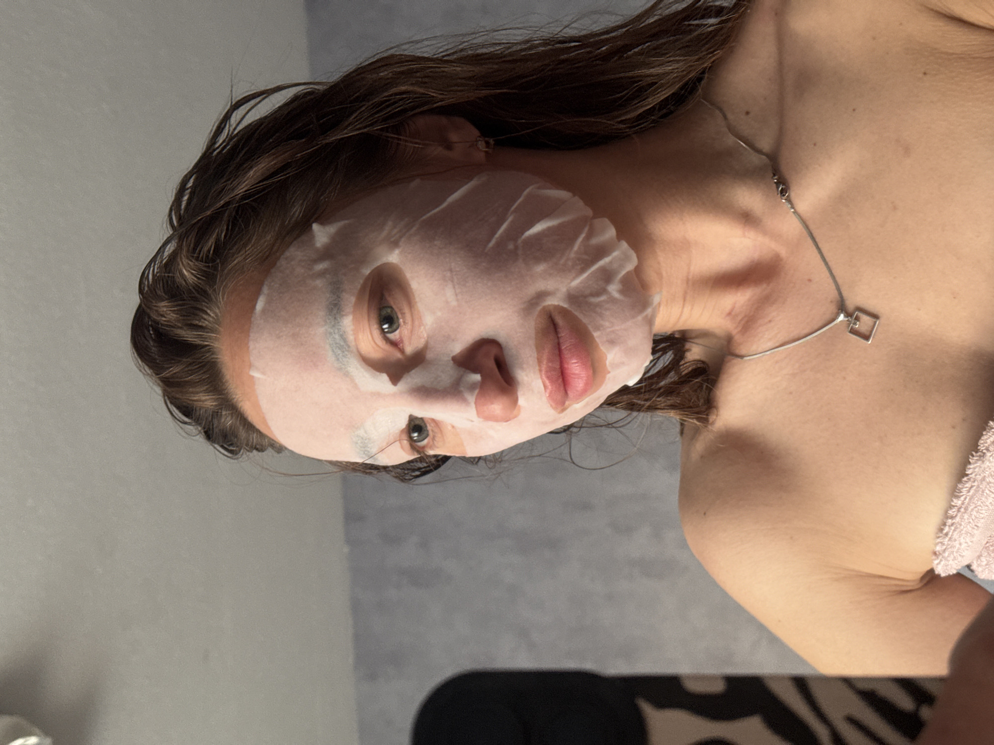 A woman with brown hair is wearing a sheet mask on her face. She is wearing a silver necklace with a square pendant. Part of her bare shoulder is visible. The environment is a room with gray walls. The image does not provide enough information to determine if this is in Chaikovsky, Russia.