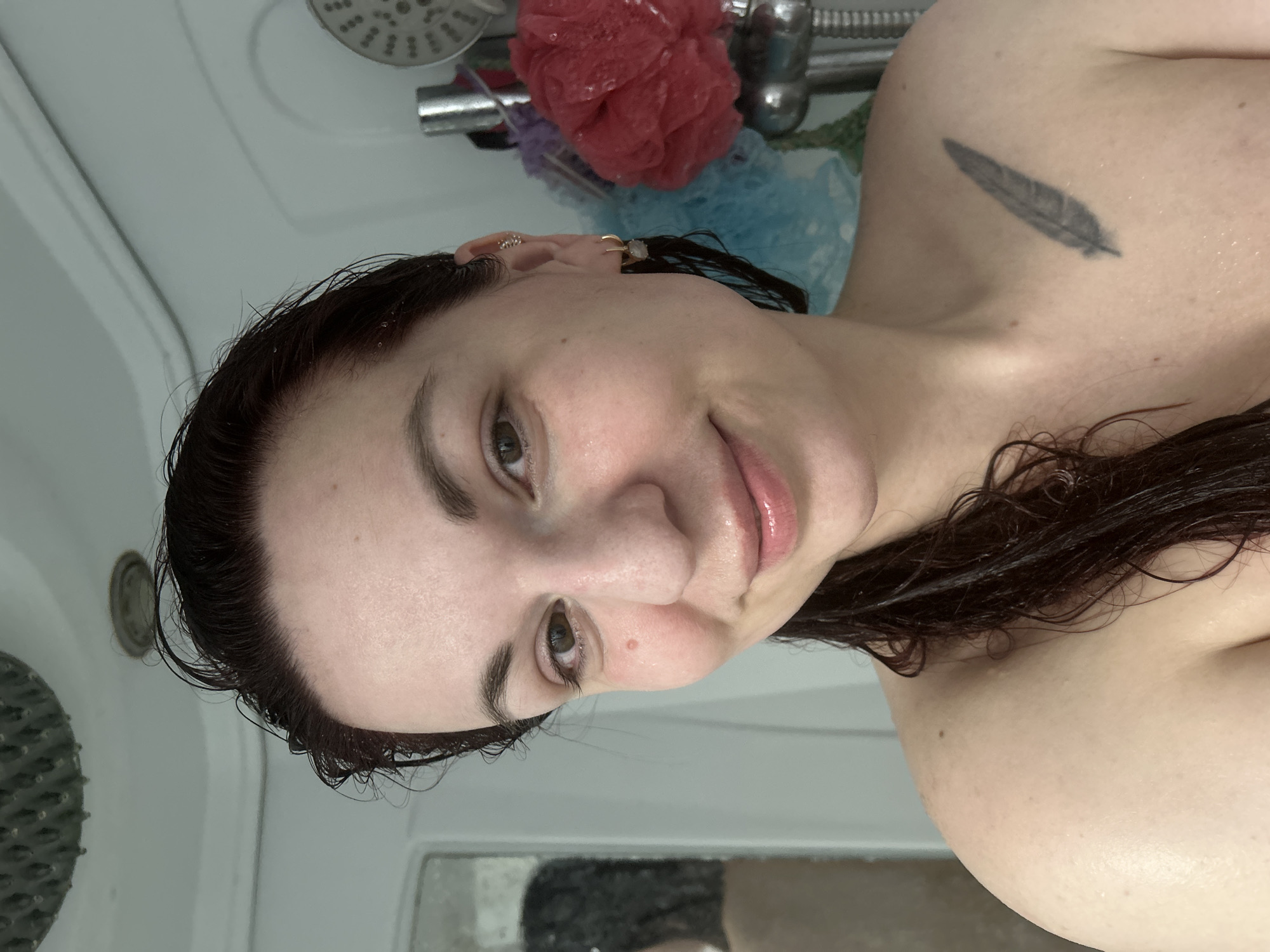 A woman is in a shower. Her hair is wet. She has a feather tattoo on her shoulder. She is looking at the camera and smiling. Behind her is a shower head and shower accessories. It is not possible to ascertain the location as the city of Фоки, Russia from the image.
