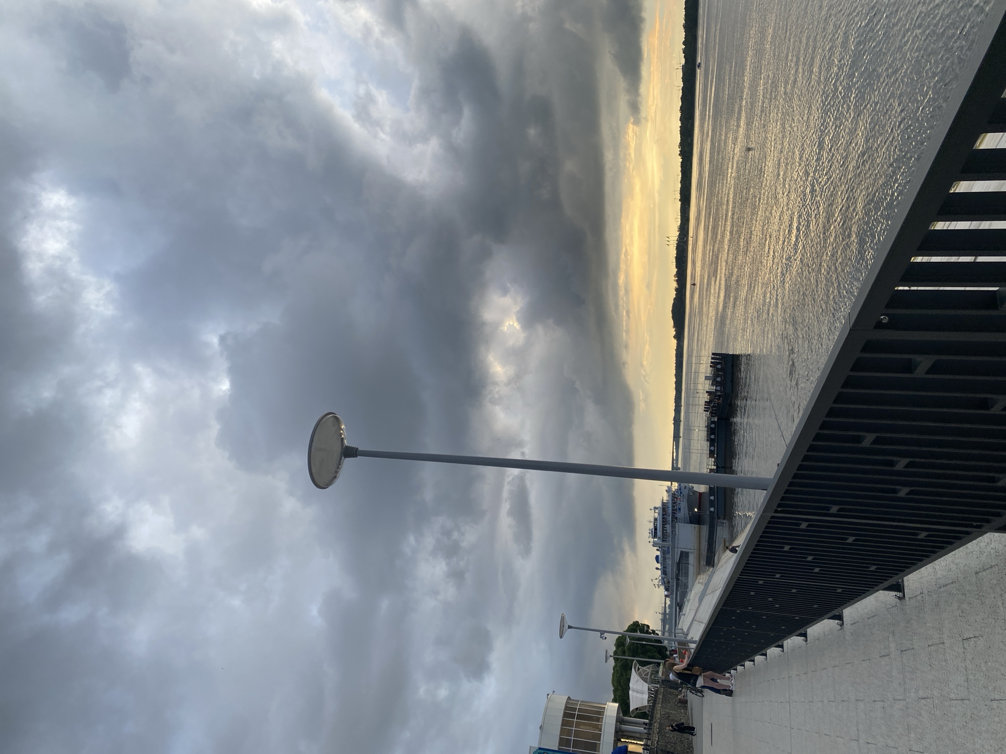 This is a vertical, eye-level, medium shot photograph taken on the embankment in the city of Perm, Russia, during the daytime, likely near sunset. The sky is dominated by dark, dramatic clouds with a sliver of golden light breaking through on the horizon over the wide, calm river. A large, white cruise ship is docked on the river, with several smaller boats and pontoons nearby.
In the foreground, a paved walkway runs along the river, bordered by a black metal railing. The walkway is lined with evenly spaced lampposts, one of which is prominently featured in the center of the frame, extending vertically towards the sky. Several people are visible on the walkway, including two women leaning on the railing and looking out at the water, and another person walking further down the path. To the left of the walkway, a modern, white building with large windows is partially visible. The river itself reflects the muted colors of the sky and the distant treeline. The overall atmosphere is serene but slightly dramatic due to the cloudy sky.