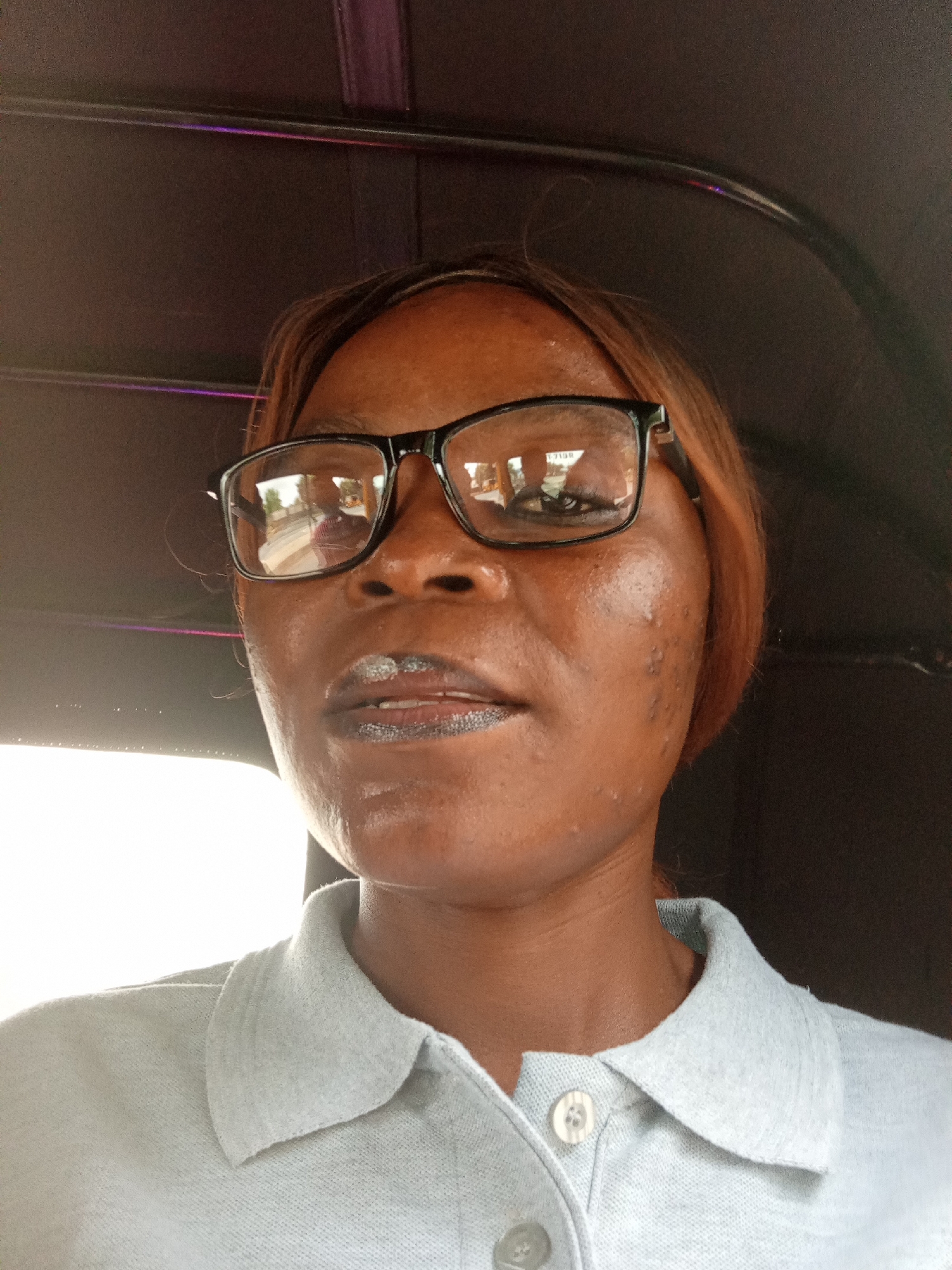 A close-up, indoor selfie features a Black woman wearing glasses and a light blue polo shirt. She is looking directly at the camera with a neutral expression. The background is dark, with hints of purple light visible at the top. The woman's skin appears slightly shiny, possibly from sweat. Her lips are colored with a shimmery silver lipstick. The scene is not identifiable as being in Zanzi, Nigeria.