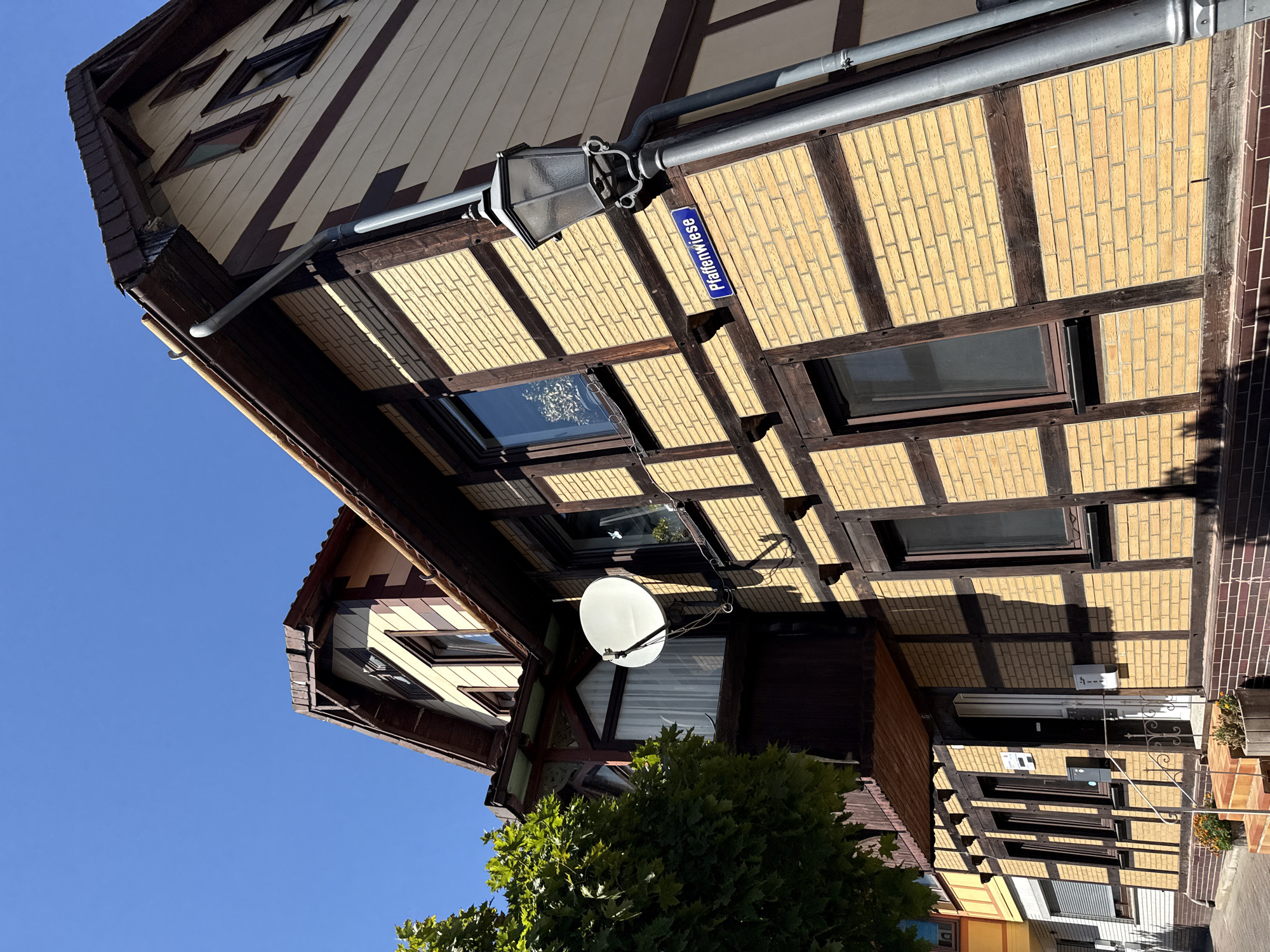 A low-angle shot captures a section of a two-story, half-timbered building with a beige and dark brown color scheme under a clear blue sky. The building's facade features yellow brickwork with dark brown wooden beams forming a diamond pattern. A dark brown roof with visible shingles is angled upwards, with a gutter system running along its edge. A white satellite dish is mounted on the wall to the left of a small balcony with white curtains visible behind a window. A streetlamp with a dark gray metal post stands to the right of the building, and a blue street sign reading "Pfaffenwiese" is attached to its pole. The lower portion of the building is visible, showing a doorway with a gray railing leading up to it, flanked by two potted plants. A large, leafy green tree is present on the left side of the frame. The scene appears to be a quiet residential or commercial street.