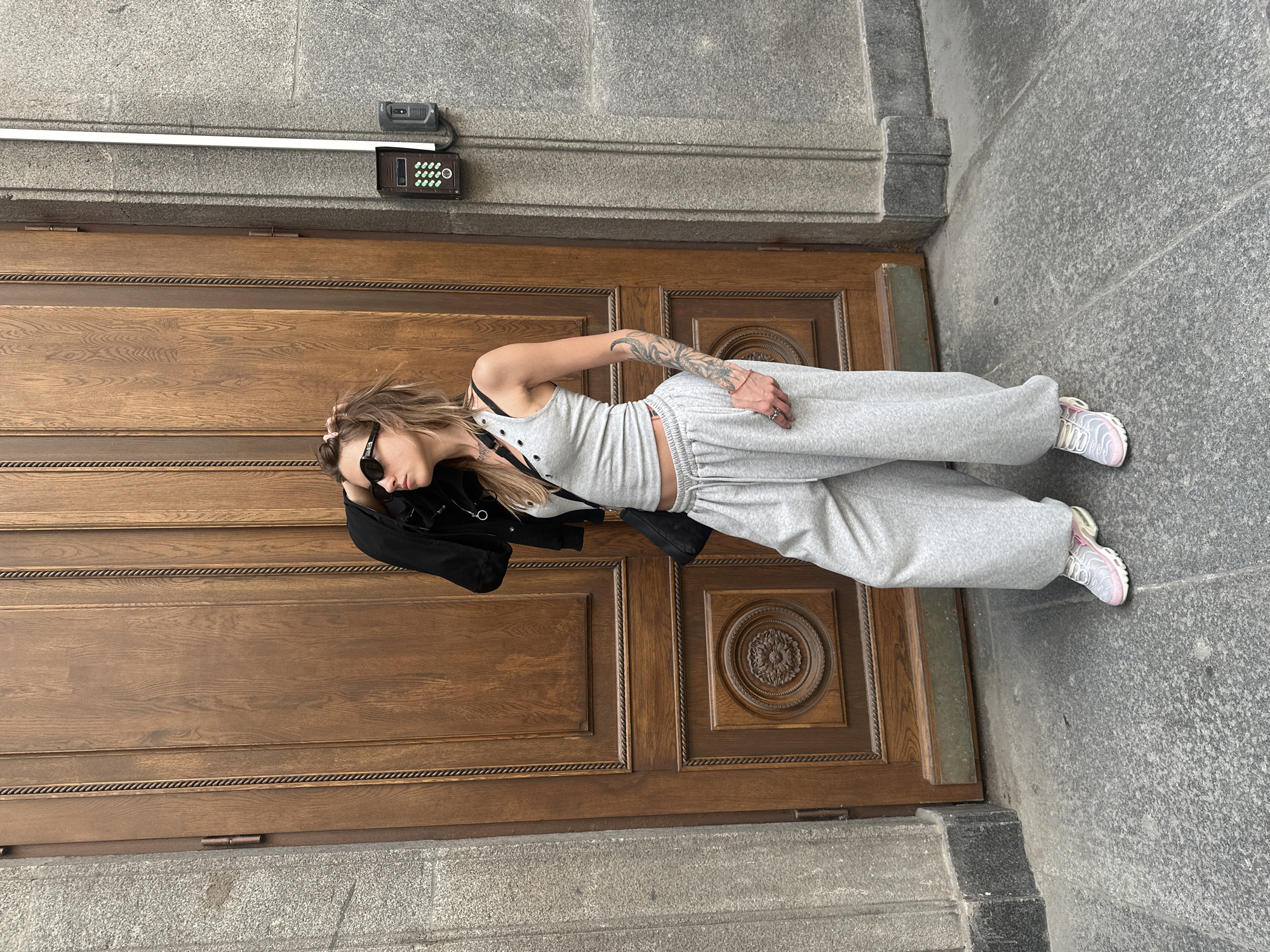 A woman stands in front of a large, ornate wooden door in an urban setting. She is wearing a grey tank top, grey sweatpants, a black jacket draped over her shoulders, sunglasses, and pink and white sneakers. She has her hand on her head, and her hair is pulled back in a ponytail. A black bag is slung across her body. The door is made of dark wood with intricate carvings. To the right of the door, there is a stone wall with a grey, textured finish. A small intercom system with a keypad and a camera is mounted on the wall. The ground in front of the door and wall is paved with grey, speckled stone. The overall impression is a candid, street-style photograph.