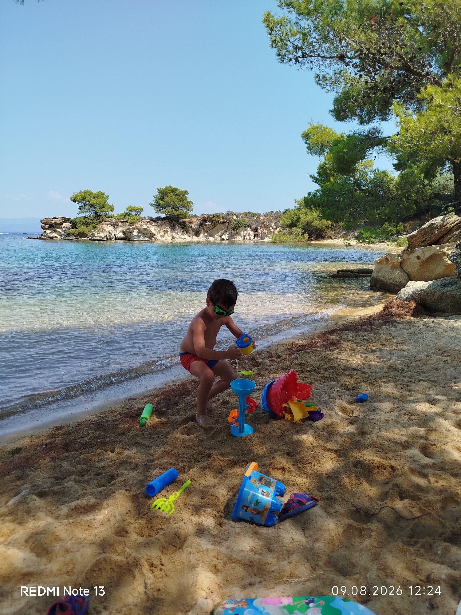 A young boy, wearing red and blue swimming trunks and green goggles, plays with a collection of colorful plastic beach toys on a sandy beach. The boy is crouched down, holding a yellow bucket and appears to be pouring water. The toys, including buckets, shovels, and sand molds, are scattered around him on the sand. The scene is set against a backdrop of clear turquoise water and a rocky coastline dotted with pine trees. The sky is a bright, cloudless blue. In the foreground, the bottom right corner of the image shows a portion of a colorful mat or towel, and the bottom left corner displays the text "REDMI Note 13". The bottom right corner displays the date "09.08.2026 12:24". This image was taken in Sithonia Municipal Unit, Greece.