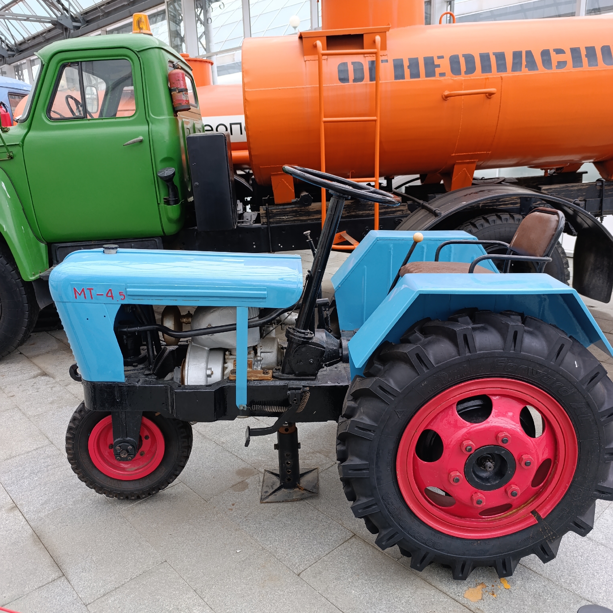 The image displays two vintage vehicles, a mini-tractor and a tanker truck, on a paved surface, likely within a museum or exhibition space in Verkhnyaya Pyshma, Russia.
In the foreground is a light blue mini-tractor, featuring striking red wheels with black, robustly treaded tires. The model designation "MT-4.5" is clearly visible in red text on its front cowling. The tractor has a black frame, a brown seat, and a black steering wheel. A mechanical jack-stand is positioned beneath its front section. Visible text on the rear tire includes "MADE IN RUSSIA" and "Forvard AC", along with tire specifications such as "93-20 B-105A".
Behind the tractor, partially obscured, is a vivid green truck cab with an amber warning light on its roof and a red fire extinguisher mounted on its side. Attached to the truck chassis is a large, bright orange tanker, which has a ladder on its side. Prominent black Cyrillic lettering on the tanker reads "ОГНЕОПАСНО" (FIRE HAZARD).
The setting is an open area with light-colored tile paving, under a large, modern roof structure with visible beams and glass panels. No people are present, and the vehicles appear to be static exhibits. The lighting is bright and even.