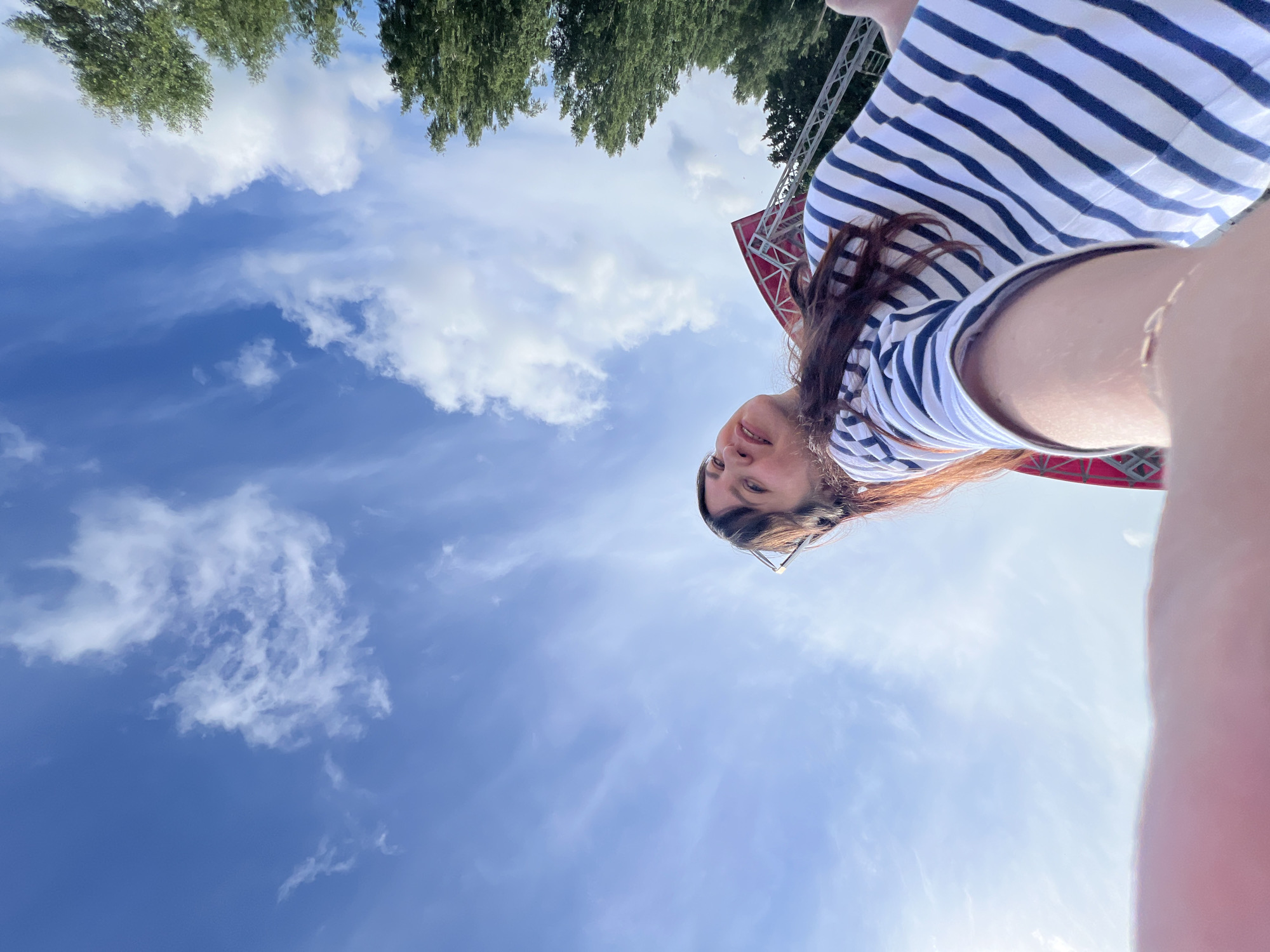 The image is a low-angle, wide-shot selfie taken outdoors under a bright blue sky with scattered white clouds. A young woman with long, dark brown hair and bangs is looking up towards the camera, holding it with her right hand, which is partially visible in the foreground. She is wearing a striped t-shirt with navy blue and white horizontal stripes. To the right of the woman, the tops of tall green trees are visible. In the background, a portion of a red and white structure, possibly a stage or scaffolding, can be seen. The lighting suggests it is daytime, likely in the late afternoon given the angle of the sun. The location is identified as Noviy, Russia.
