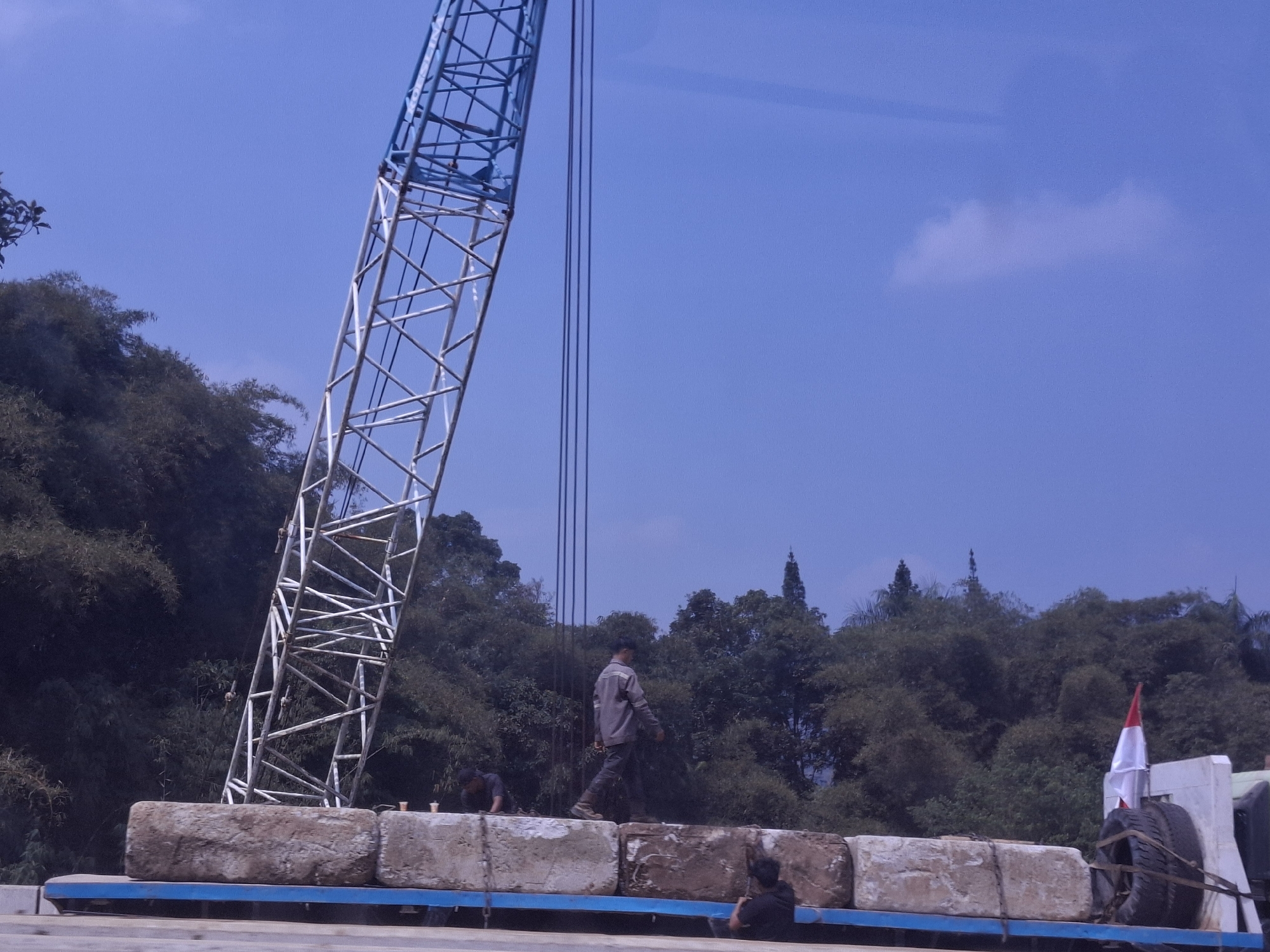 The image captures a construction scene under a clear blue sky with scattered clouds. A large blue and white metal crane dominates the left side of the frame, its latticed structure extending upwards. In the foreground, a flatbed trailer is loaded with several large, rough-hewn concrete blocks.
Three individuals are visible. One person, wearing a grey jacket and dark pants, walks along the top of the concrete blocks. Another person, in a dark shirt, is crouched down on the blocks near the left side of the trailer. A third person, partially visible from behind and wearing a dark hoodie, is seated on the right side of the trailer.
Behind the trailer and the construction equipment, a dense line of lush green trees forms a backdrop, suggesting a location with abundant vegetation, possibly on the outskirts of a built-up area. To the far right, a white truck is partially visible, with a spare tire and a red and white Indonesian flag mounted on it. The overall impression is one of ongoing construction or material transport in a semi-rural or natural setting. This scene is consistent with typical construction activities that might take place in areas such as Karangtengah, Indonesia, where such landscaping and development are common.