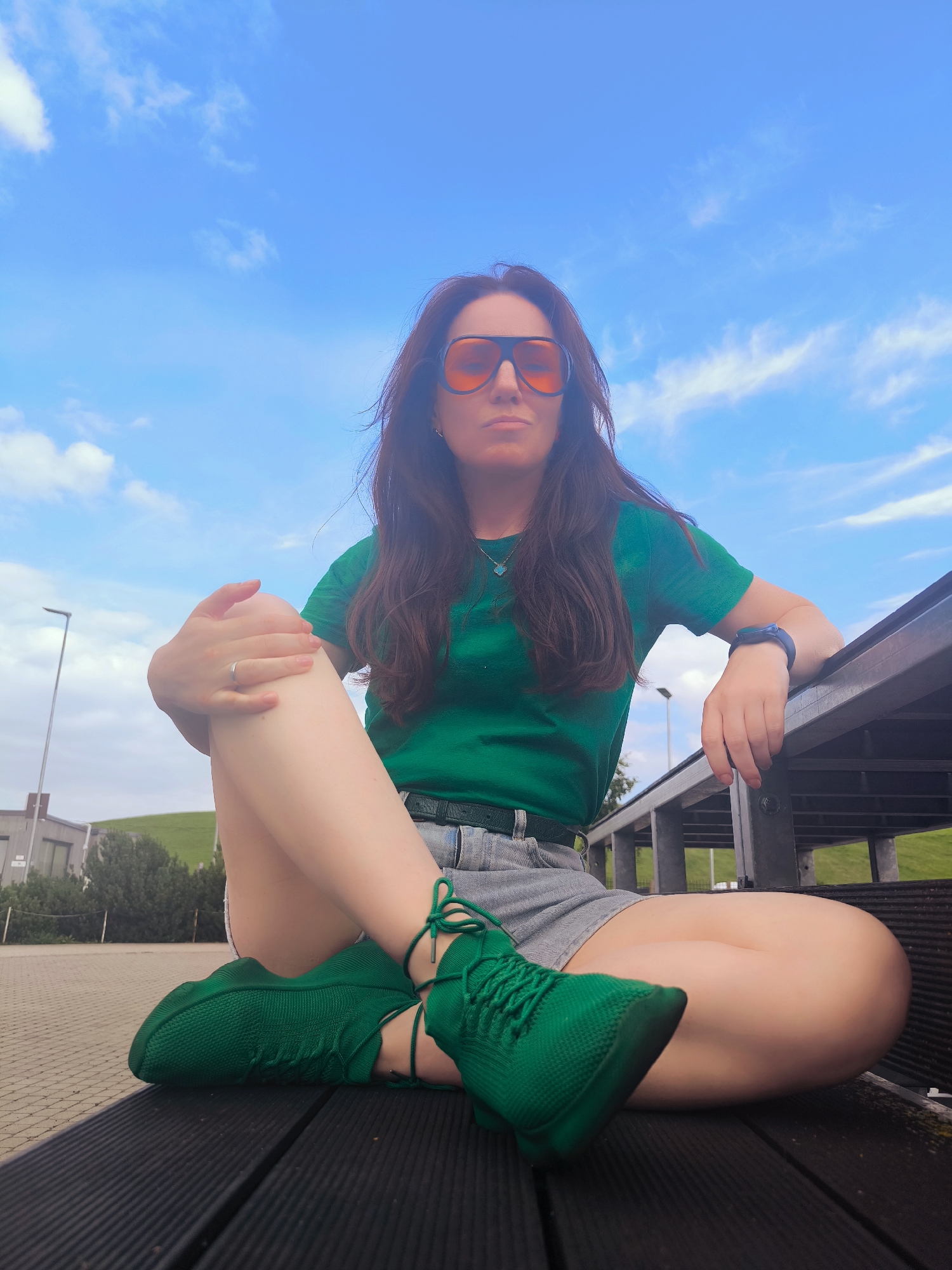 A woman is sitting on a dark wooden bench outdoors, with her legs crossed. She is wearing a green t-shirt, grey denim shorts, and bright green sneakers. She has on large orange-tinted sunglasses and a silver ring on her left hand. A dark smartwatch is visible on her right wrist. The background shows a bright blue sky with scattered white clouds, some buildings, and a grassy hill. The overall impression is casual and summery. The location is the Vērmanes Garden in Riga, Latvia.