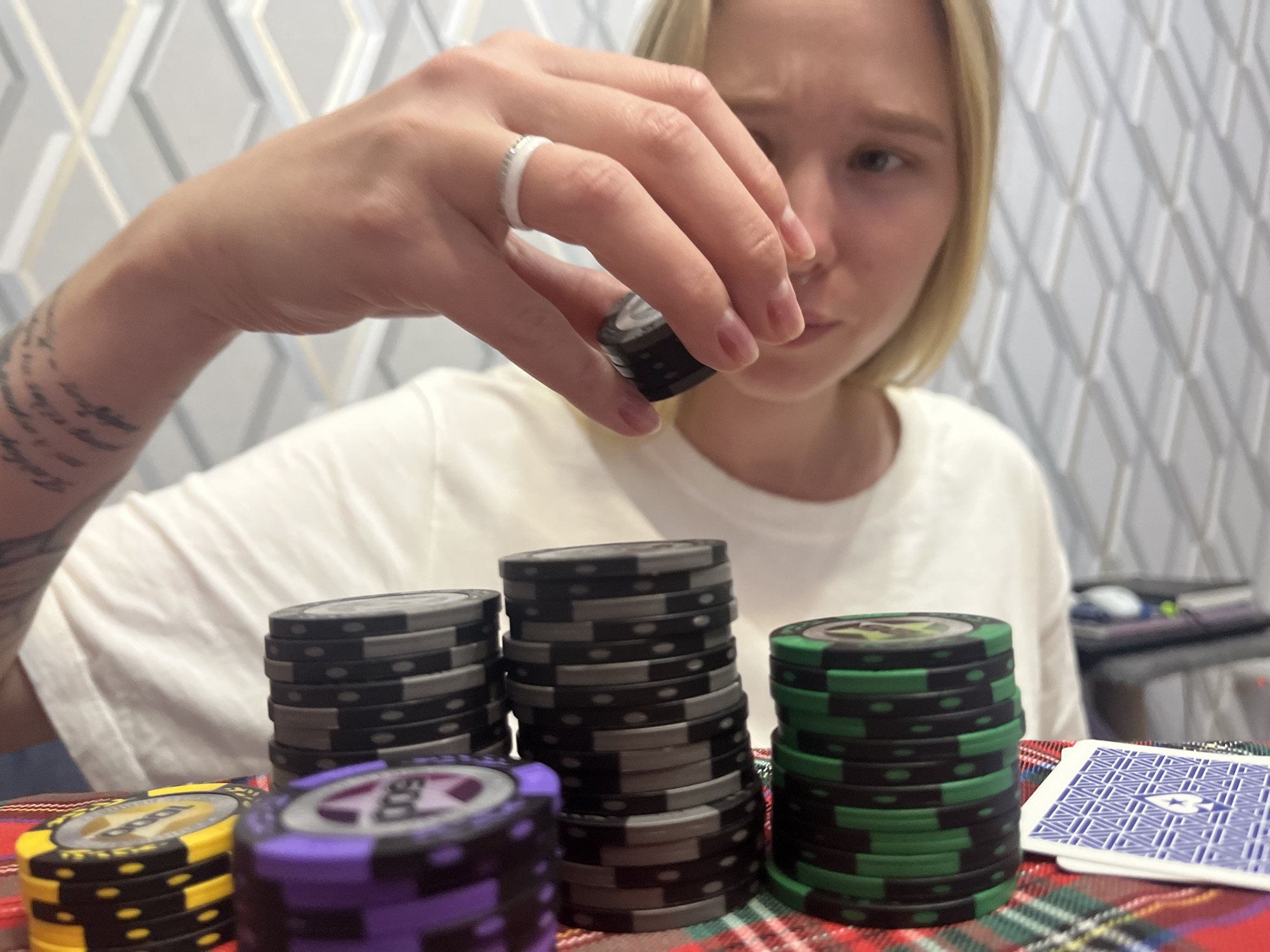 A young woman with blonde hair, wearing a white t-shirt and a silver ring on her left hand, is holding a black poker chip between her thumb and forefinger, bringing it closer to her face. She has a tattoo on her left forearm. In front of her, on a red and green plaid surface, are several stacks of poker chips: one stack of yellow and black chips, one stack of purple and black chips with "500" visible, a tall stack of black and gray chips, and a stack of green and black chips. Two playing cards with a blue and white geometric pattern are also visible on the right side of the image. The background features a textured, diamond-patterned wallpaper. The scene appears to be indoors. There is no identifiable information to determine the location in Budva, Montenegro.