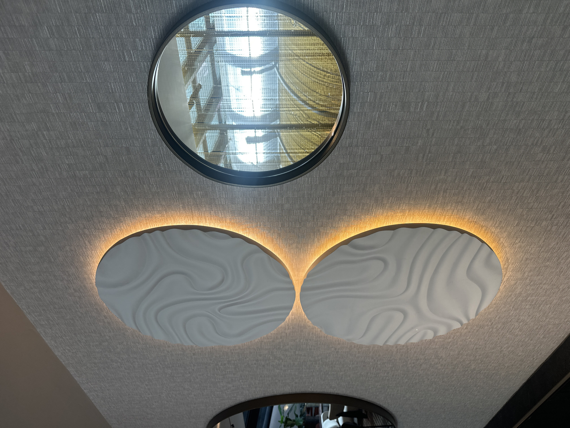 The image shows a textured wall with two circular, white relief sculptures on the left, each illuminated from behind by a warm orange light. To the right of the sculptures is a round mirror with a dark frame. The mirror reflects a bright, open space with what appears to be a ceiling structure composed of beams and mesh, and a yellow, textured floor or surface below. Another circular mirror with a dark frame is partially visible on the far left of the image, reflecting a room interior with furniture and a potted plant. The overall scene suggests a modern interior design, possibly in a public space or a well-decorated home. The location is unknown.