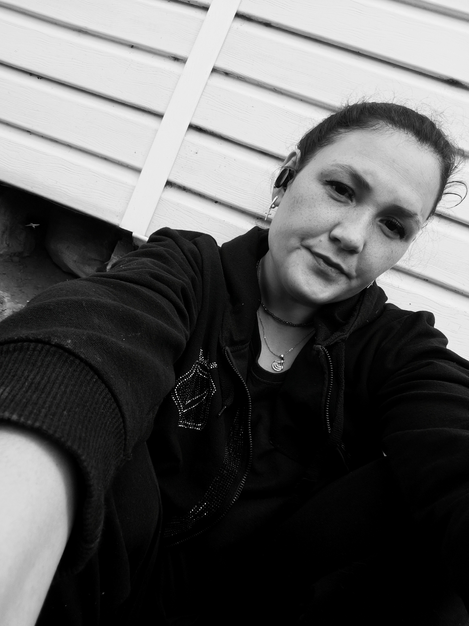 A black and white, medium-shot, selfie-style photograph shows a woman with her hair pulled back, wearing a black zip-up hoodie over a black t-shirt and a silver necklace with a crescent moon charm. She has a small cross earring in her left ear and a dark earpiece in her right ear. Her left arm is visible in the foreground, bent at the elbow and resting on her leg. The background consists of white horizontal siding, suggesting an outdoor setting, possibly a residential exterior. There are no identifiable landmarks that would place this image in Karsha, Russia.