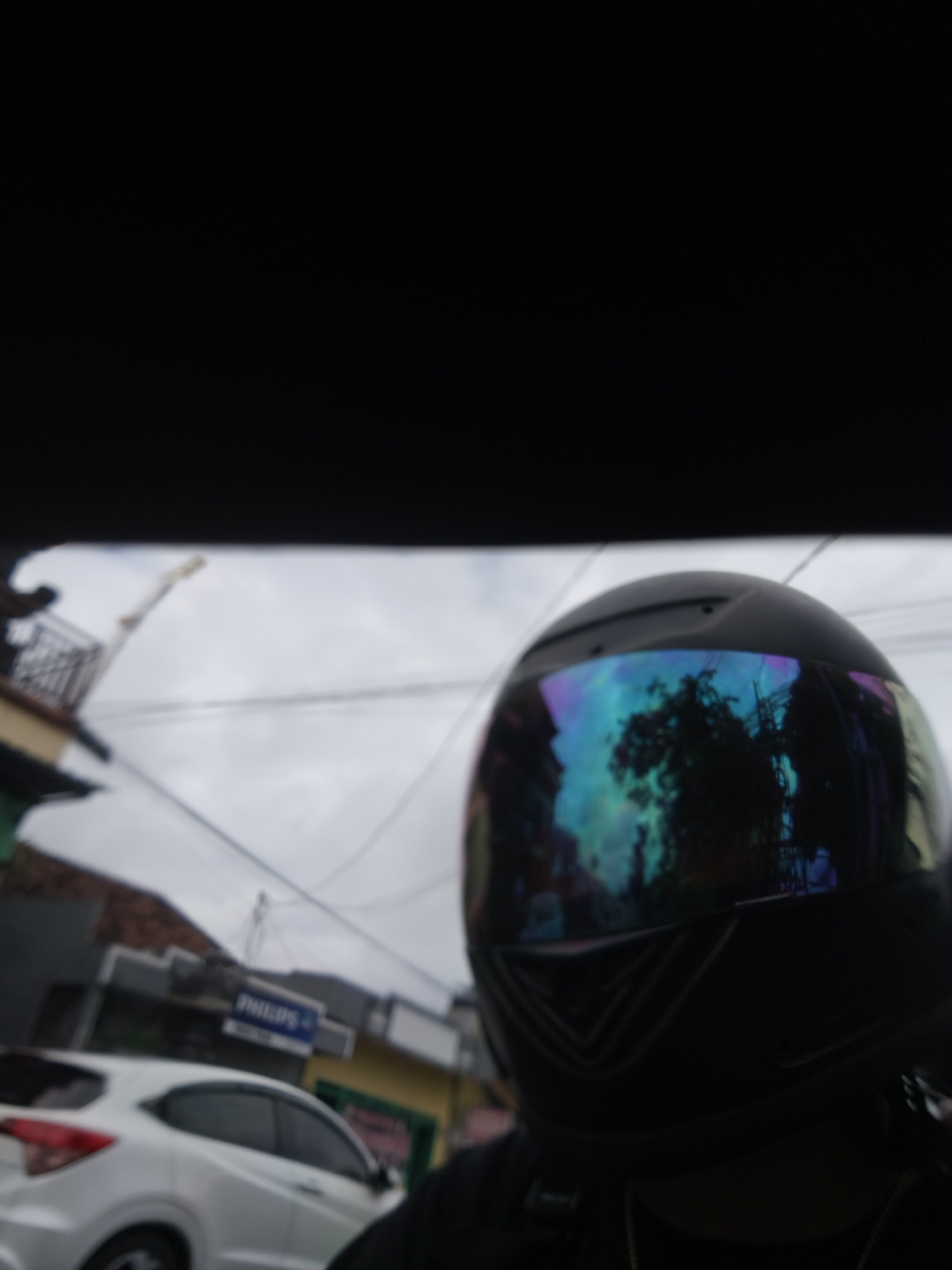 The image shows a close-up of a person wearing a black motorcycle helmet with a reflective visor. The visor displays a colorful, iridescent reflection of trees and buildings, suggesting an outdoor environment. In the background, partially blurred, are street scenes of Denpasar, Indonesia. A white car is visible on the left, and buildings with signage, including one that reads "PHILIPS," line the street. Overhead, power lines crisscross the sky, which is overcast. The overall impression is a snapshot taken from the perspective of someone riding a motorcycle through a city.