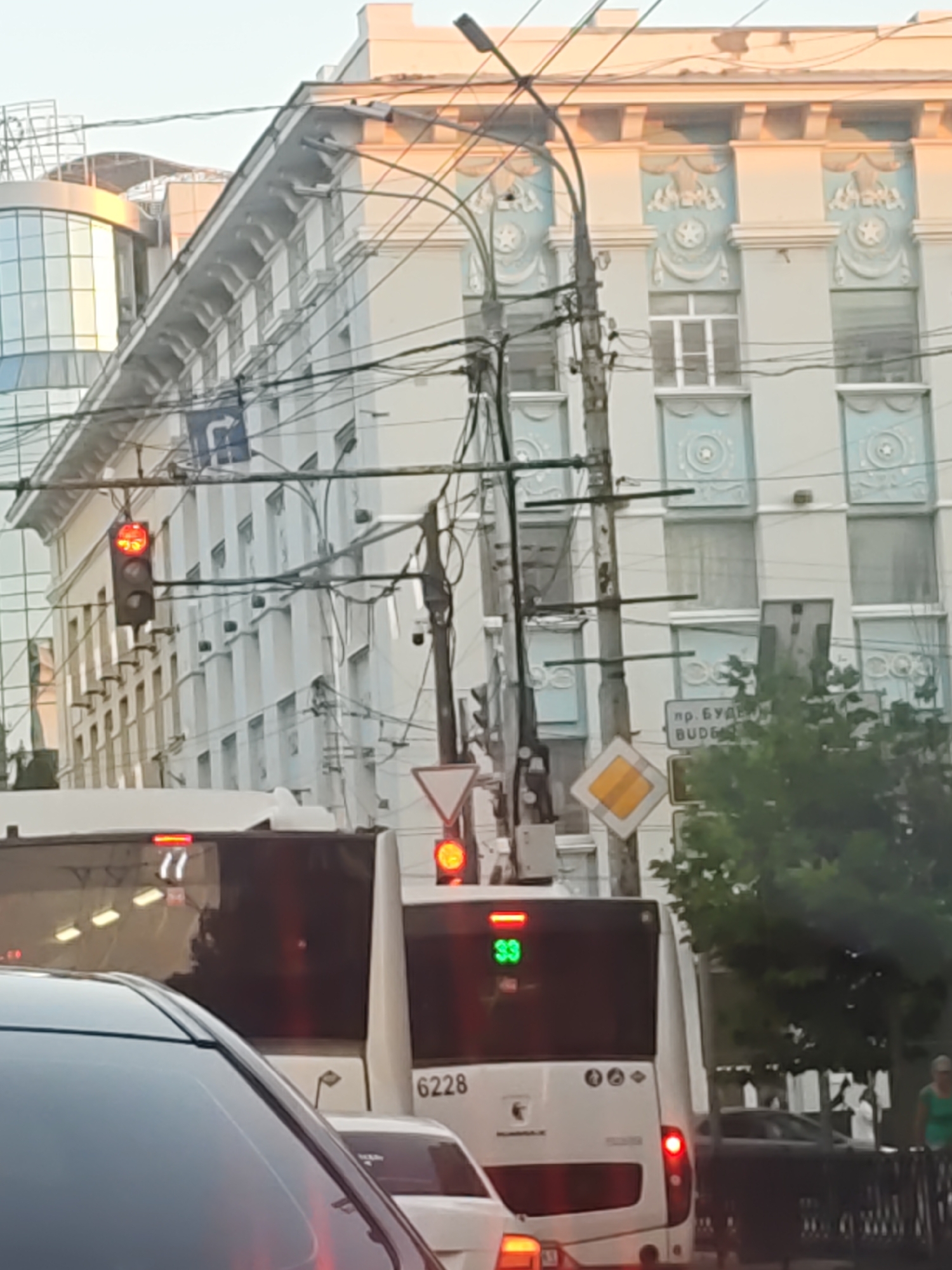 The image depicts a street scene in Rostov-on-Don, Russia, likely during daylight given the illumination. In the foreground, the rear of a white bus with the number "6228" is visible, along with its license plate digits which are partially obscured but appear to end in "77". To the left, another white bus is partially seen. Multiple traffic lights are present, one showing red. There are also street signs and a diamond-shaped yellow sign. Overhead, a complex network of wires, likely for trolleybuses or trams, crisscrosses the sky. Behind the buses, a multi-story building with ornate architectural details in light blue and white is prominent, featuring decorative elements around the windows. Further back, another modern building with glass facades can be seen. Trees and greenery are present on the right side of the frame, and a person in a green shirt is walking in the background. The scene suggests a busy urban environment with active traffic.