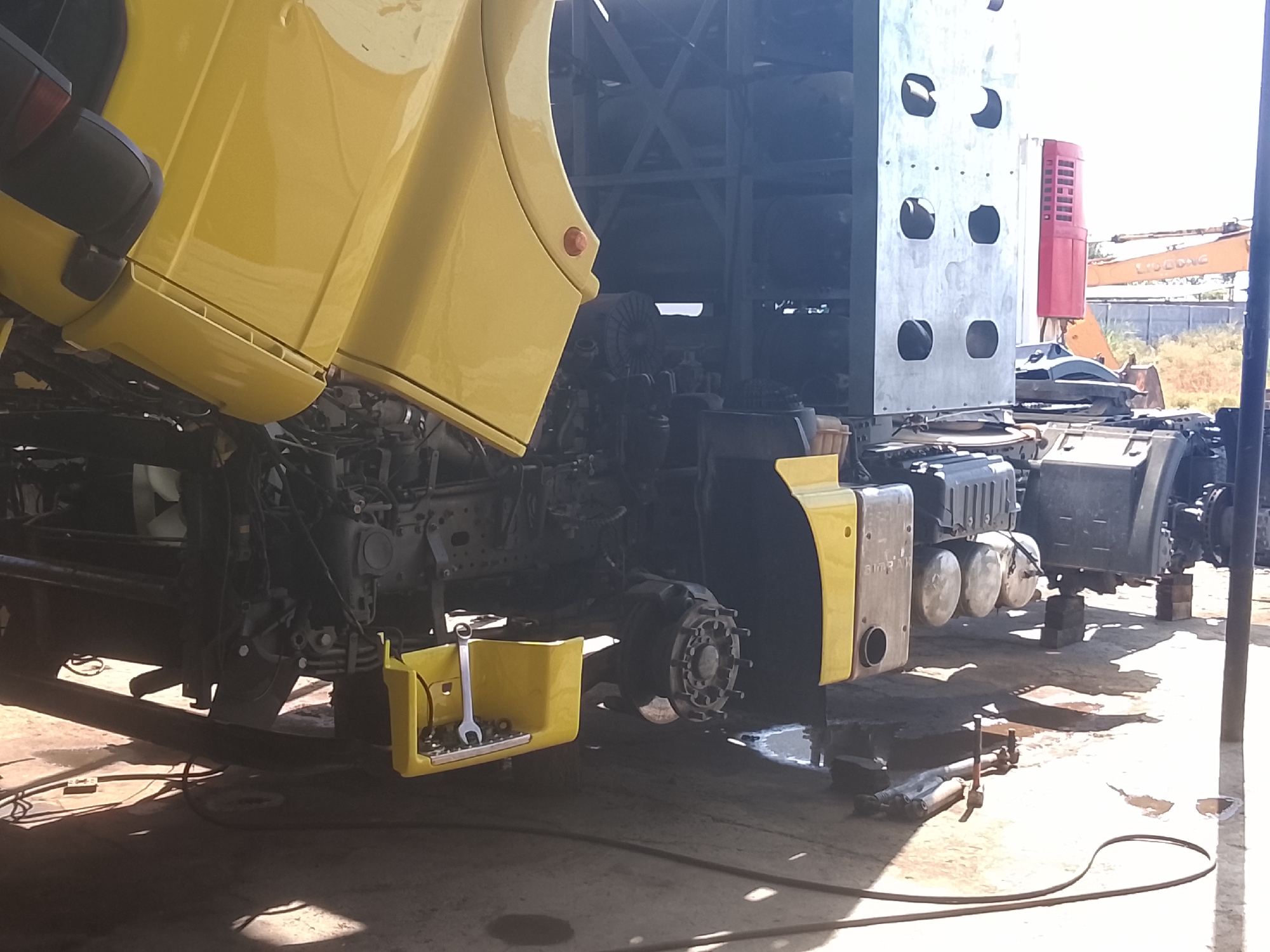 In Sabir Rakhimov, Uzbekistan, a yellow truck cab is tilted upwards, revealing the engine and chassis. The truck is undergoing maintenance, with its front wheel removed and its chassis supported by blocks. A yellow toolbox sits on the ground near the front wheel, containing various tools, including a wrench. In the background, other vehicles and construction equipment are visible, suggesting a repair shop or industrial area. The scene is outdoors, with bright sunlight casting shadows on the concrete ground.