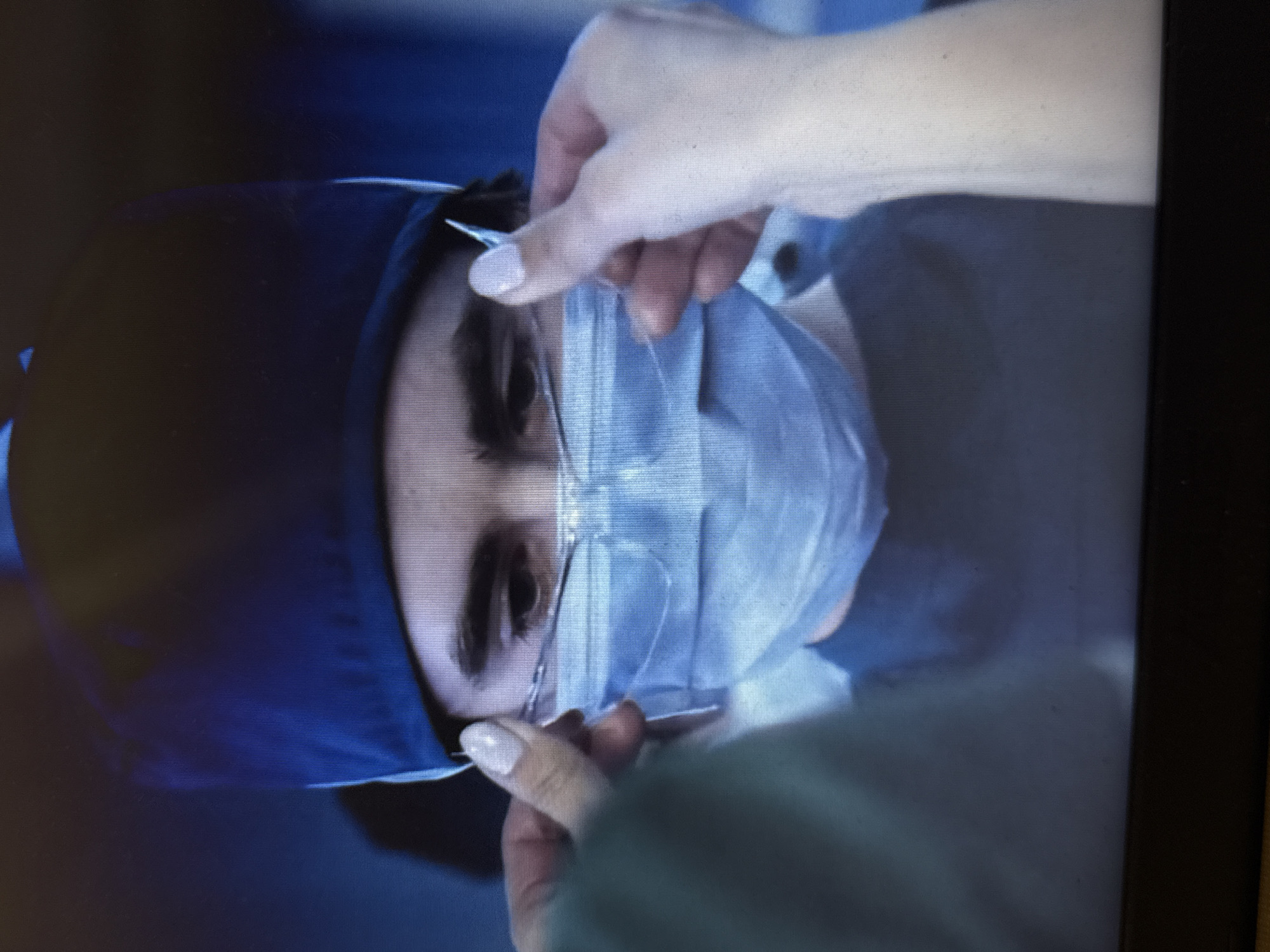 The image shows a close-up of a person wearing a blue surgical cap and a light blue surgical mask. The person has dark eyebrows and is looking forward. Their eyes are visible above the mask. A hand, with a light-colored nail, is adjusting a pair of clear glasses over the mask. Another hand, partially visible on the left, is also involved in this action. The background is dark and out of focus, with some blue tones suggesting an indoor, possibly medical, setting. The image appears to be a still frame from a video or television program. The location is unknown.